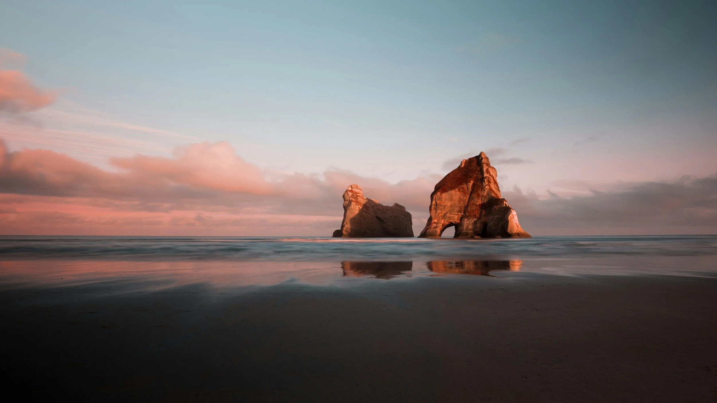 A seascape with two large rocky formations in the water at sunset or sunrise, pink and blue hues in the sky, and calm waves reflecting the rocks on a sandy beach in the foreground.