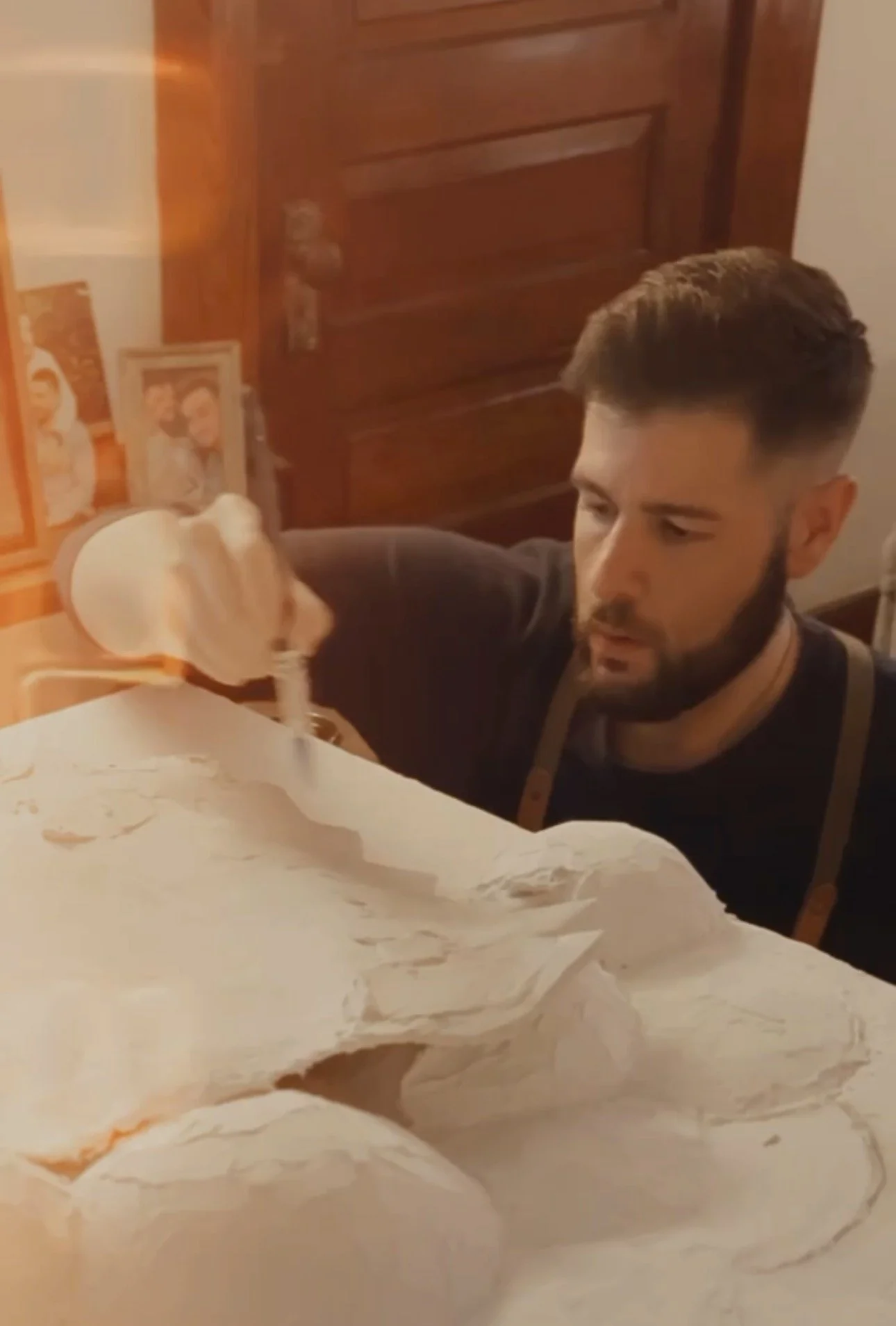 A man with a beard is pouring milk from a bottle onto a large piece of bread on a table, with family photos visible in the background.