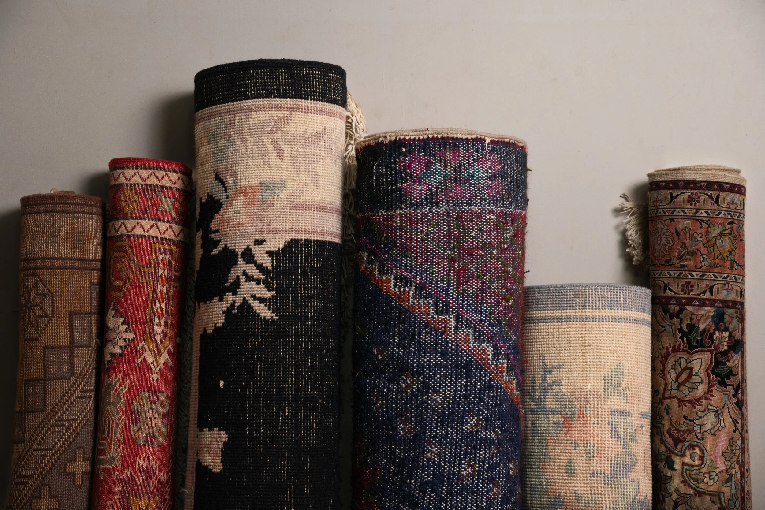 A series of rolled woven rugs with various intricate designs and colors, standing upright against a plain white wall.