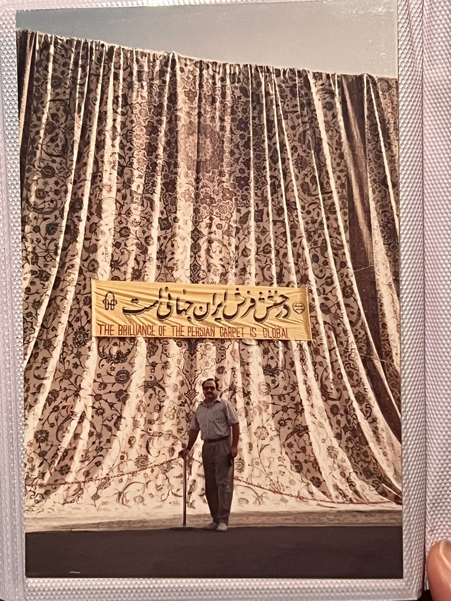A man in a shirt and pants standing in front of large ornate curtains with a sign that reads 'The brilliance of the Persian carpet is global' written on it in both English and Persian.