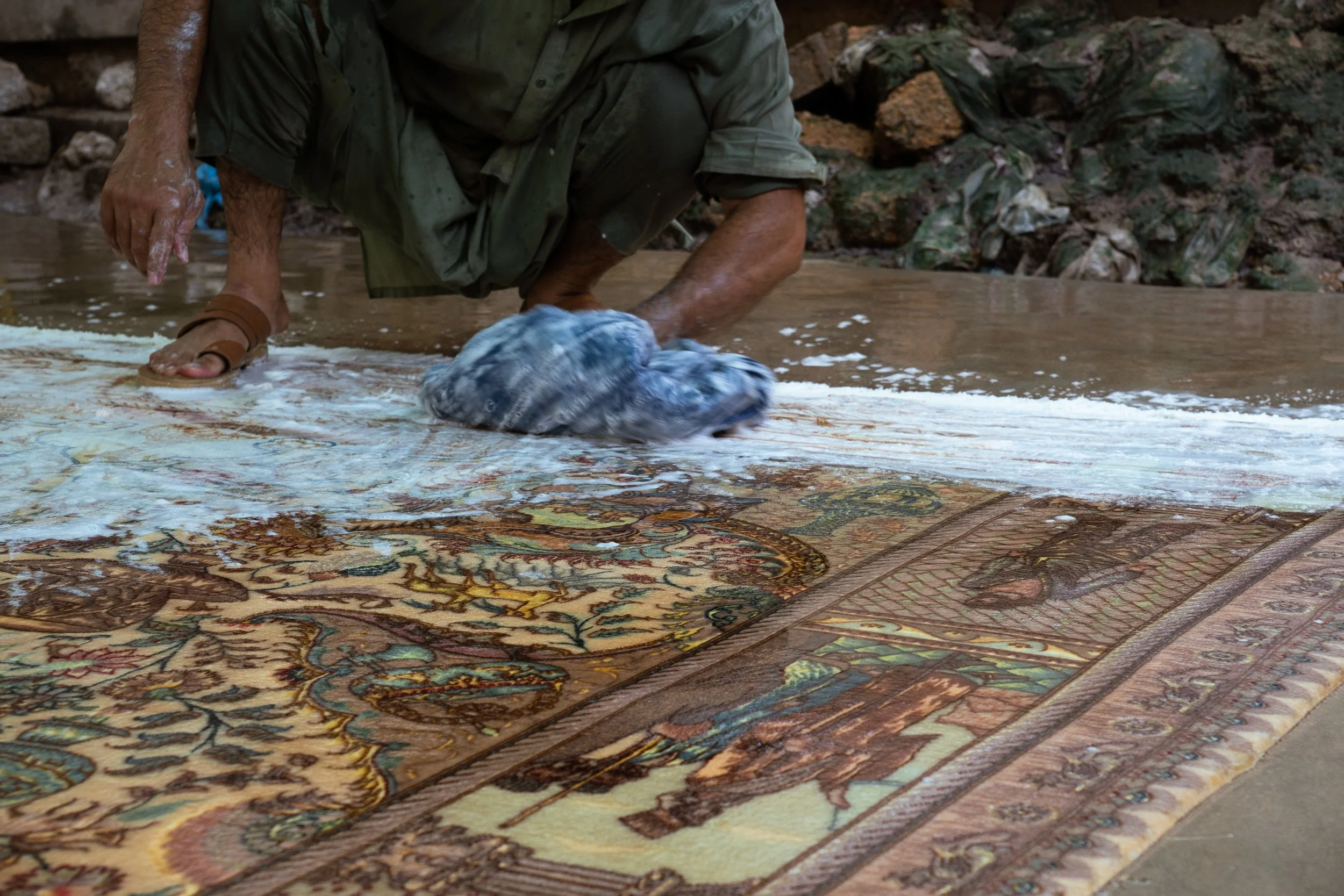 Person crouching in water, cleaning a large traditional patterned rug outdoors, with rocks and green foliage in the background.