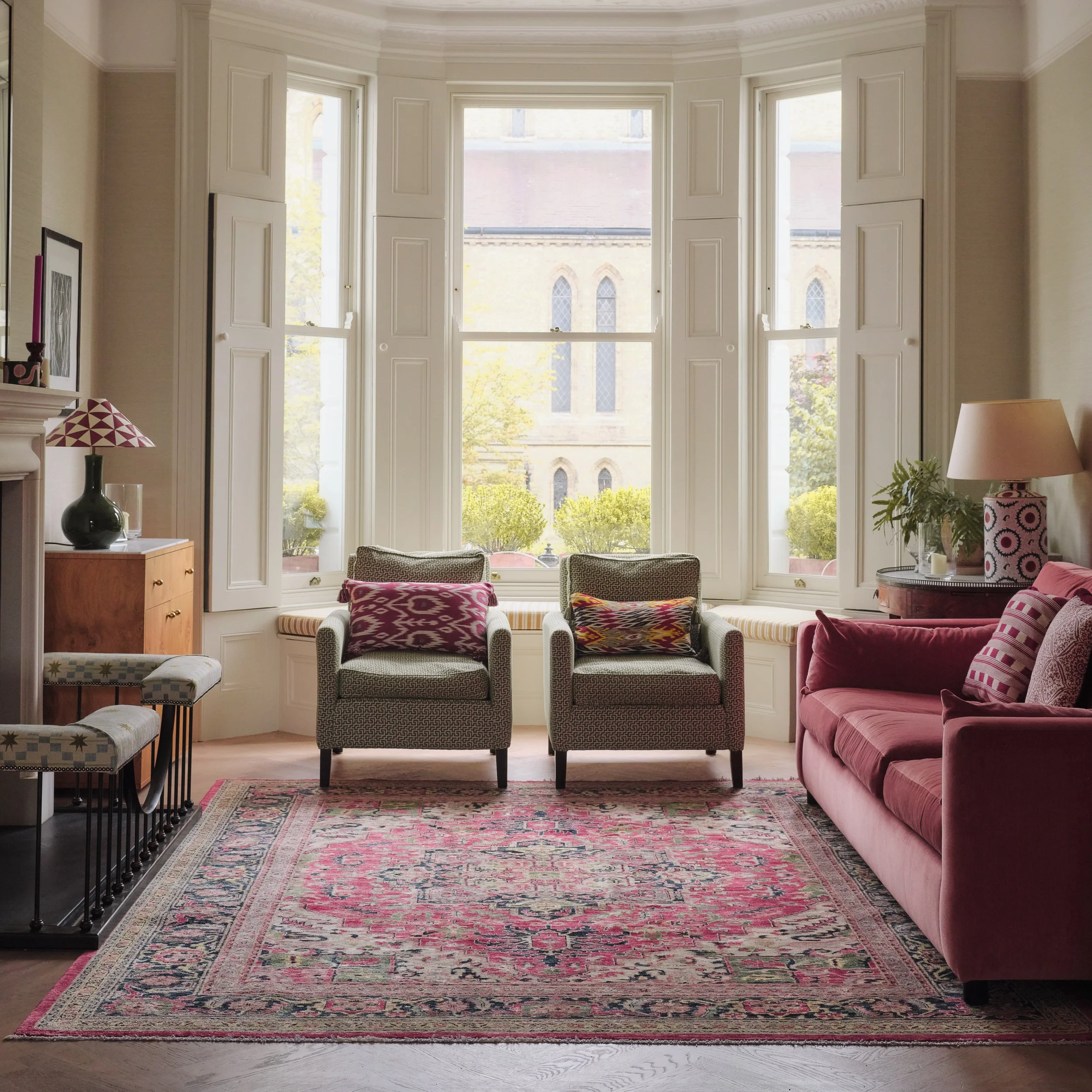 Living room with large bay window, patterned area rug, pink velvet sofa, armchairs with patterned upholstery, side table with lamp, decorative objects, and framed pictures on the wall