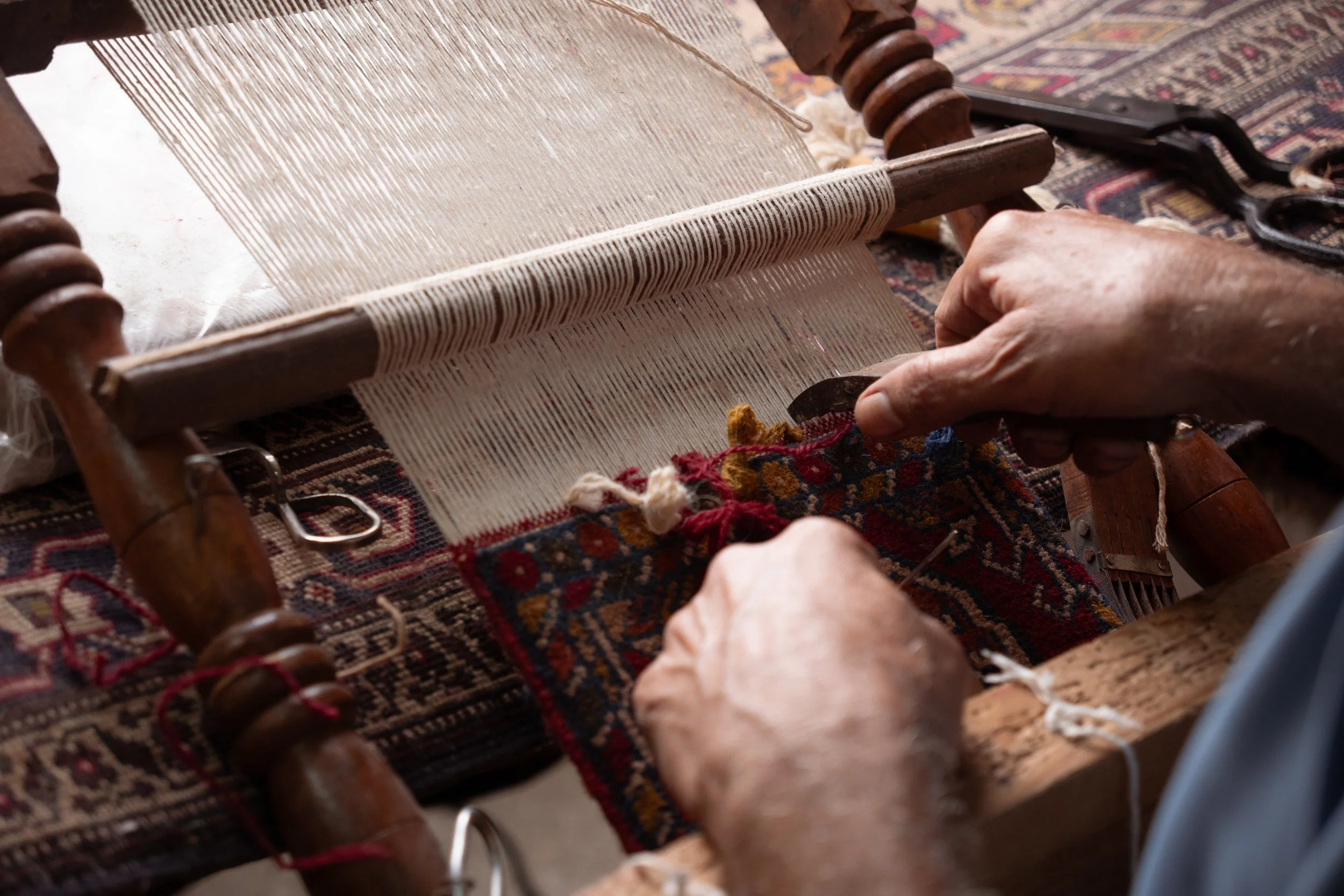 A person is weaving fabric on a wooden loom, with various threads and yarns visible, on an ornate patterned rug.
