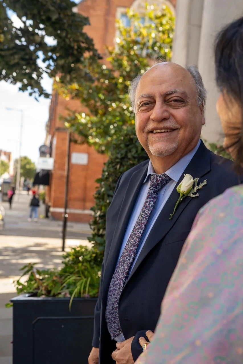 Smiling elderly man in a suit and tie with a white flower boutonniere, outdoors on a sunny day, engaged in conversation with a woman whose back is to the camera.