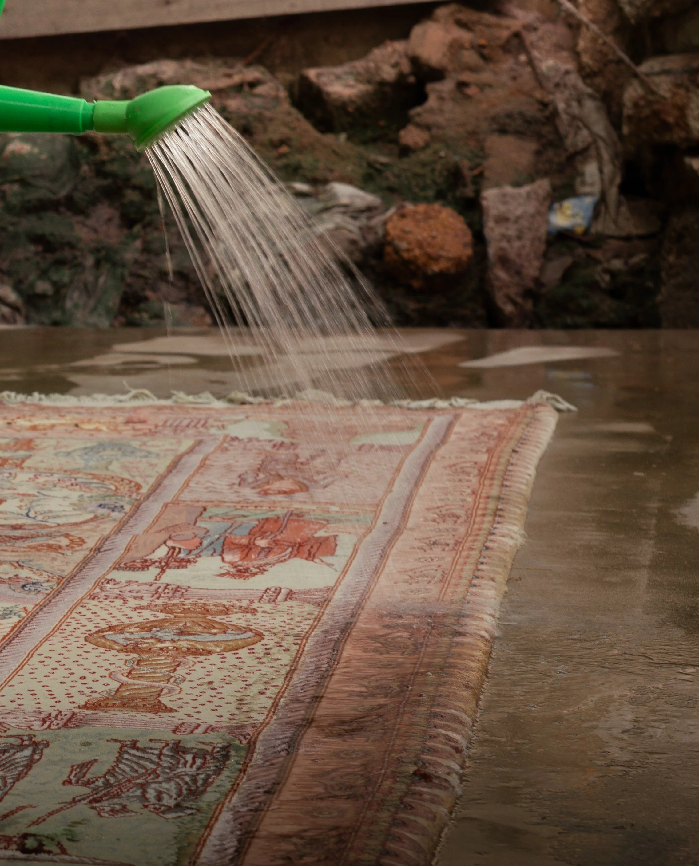 A green garden watering can pouring water onto an ornate Persian-style rug on the floor, with a rocky and earthy background.