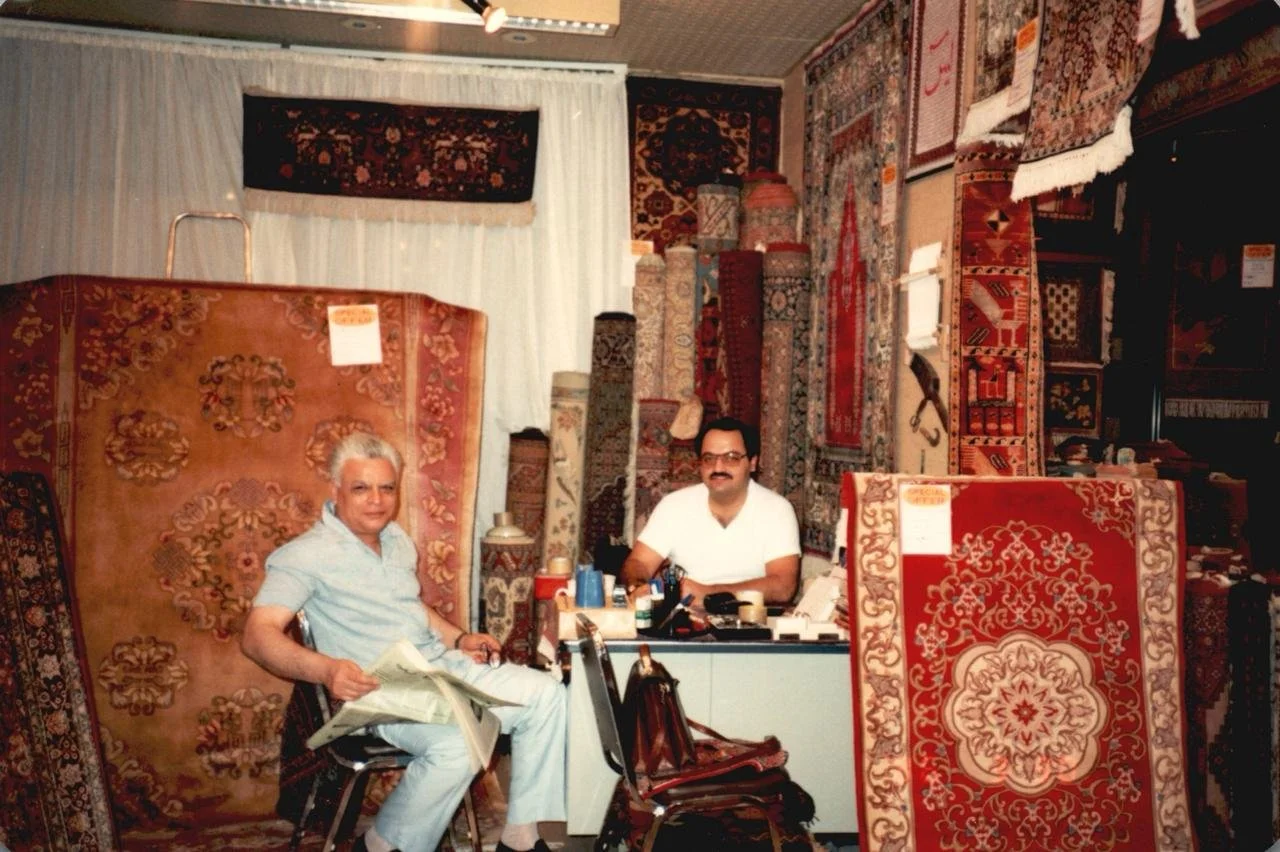 Two men sitting in a room filled with hanging and rolled-up Persian rugs and carpets.
