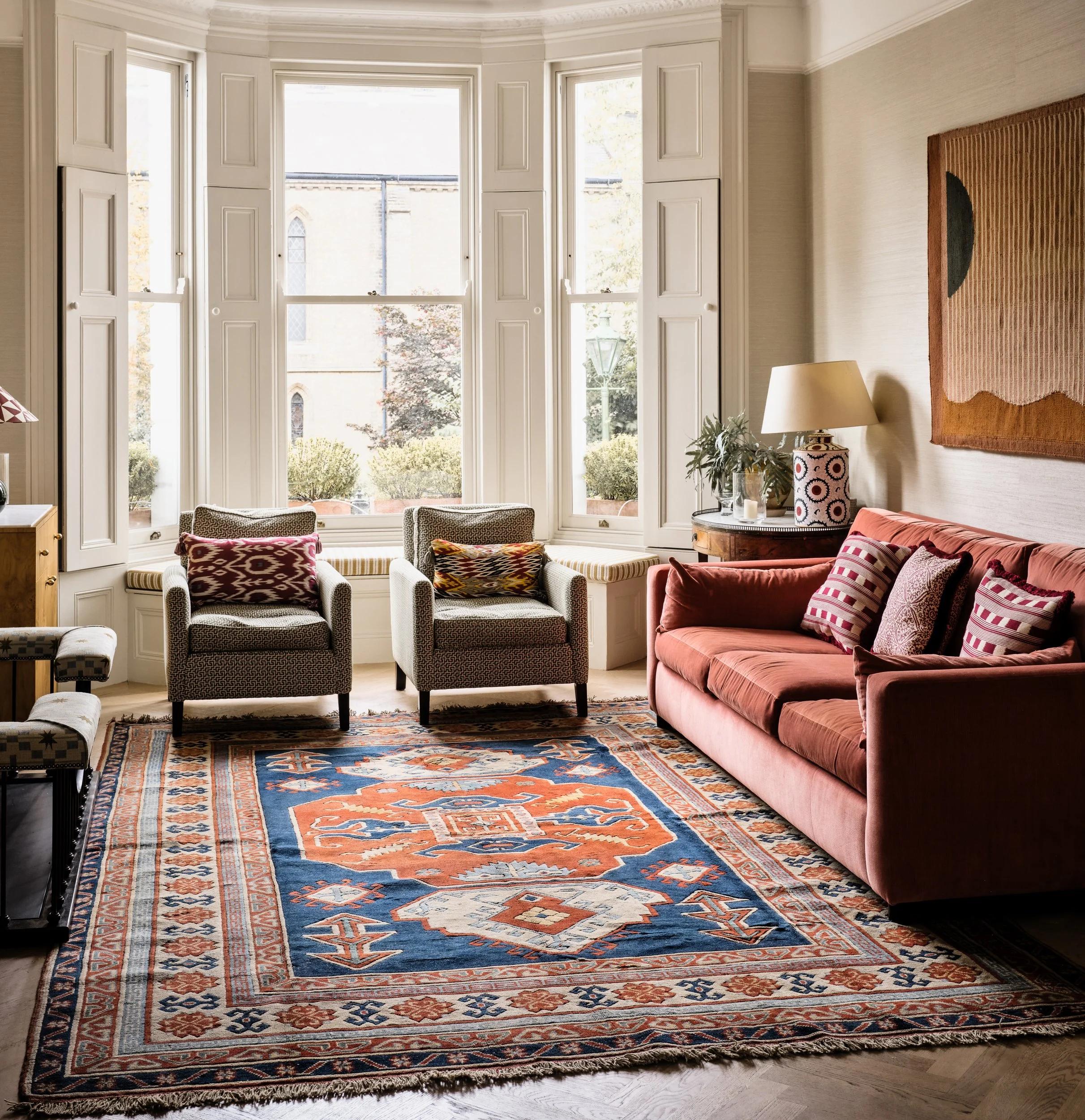 A living room with a colorful oriental rug, a coral sofa with cushions, two patterned armchairs, a side table with a lamp, and a large window with white trim showing a view of outdoor trees and a building.