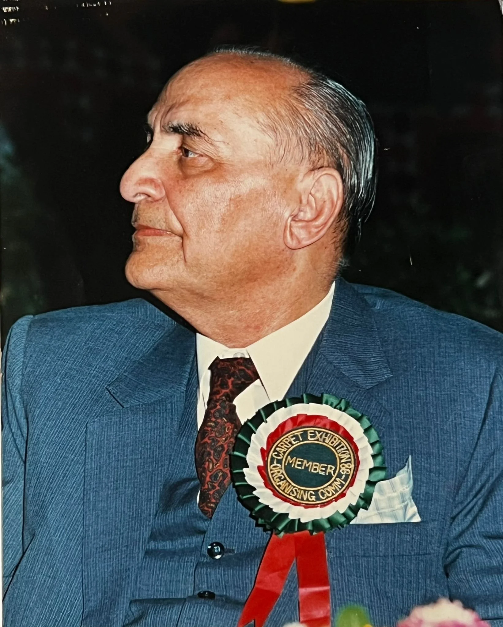 Side profile of an older man with gray hair, wearing a blue suit, white shirt, and a tie, with a large rosette ribbon badge on his chest that reads 'Club Carpet Exhibition Member'.