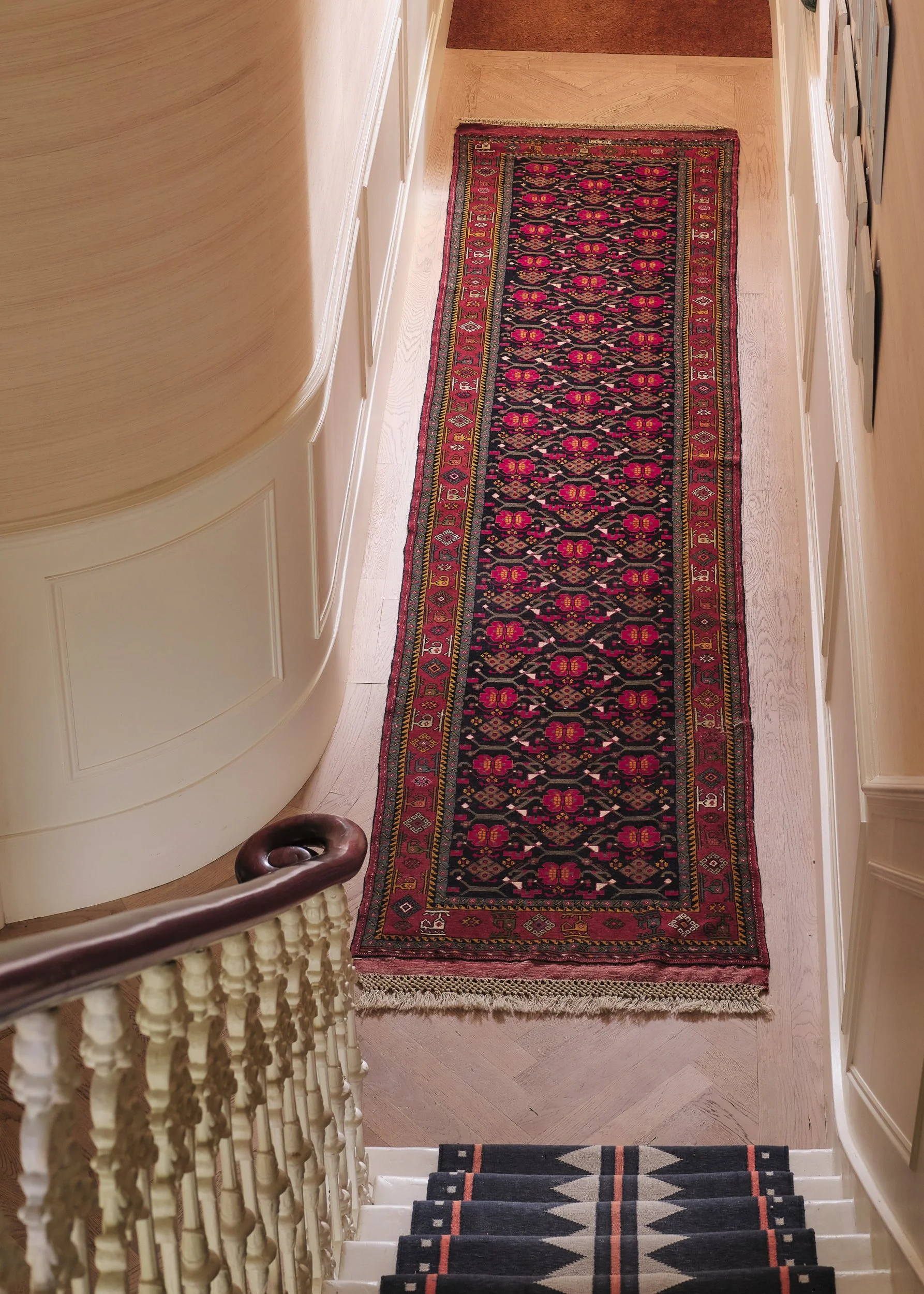 View looking down a staircase with a patterned black and gray runner rug, wood floors, and cream-colored walls with white trim. A red and pink patterned runner rug is on the adjacent hallway floor.