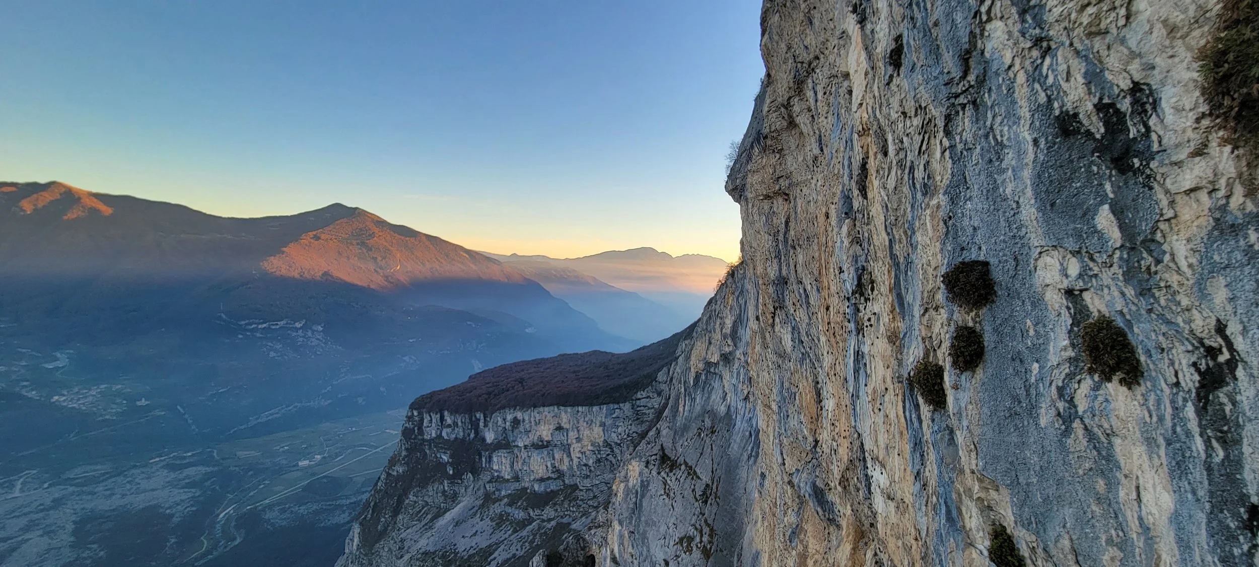 Blick auf eine Berglandschaft bei Sonnenaufgang, mit steiler Felswand im Vordergrund und Tälern im Hintergrund.