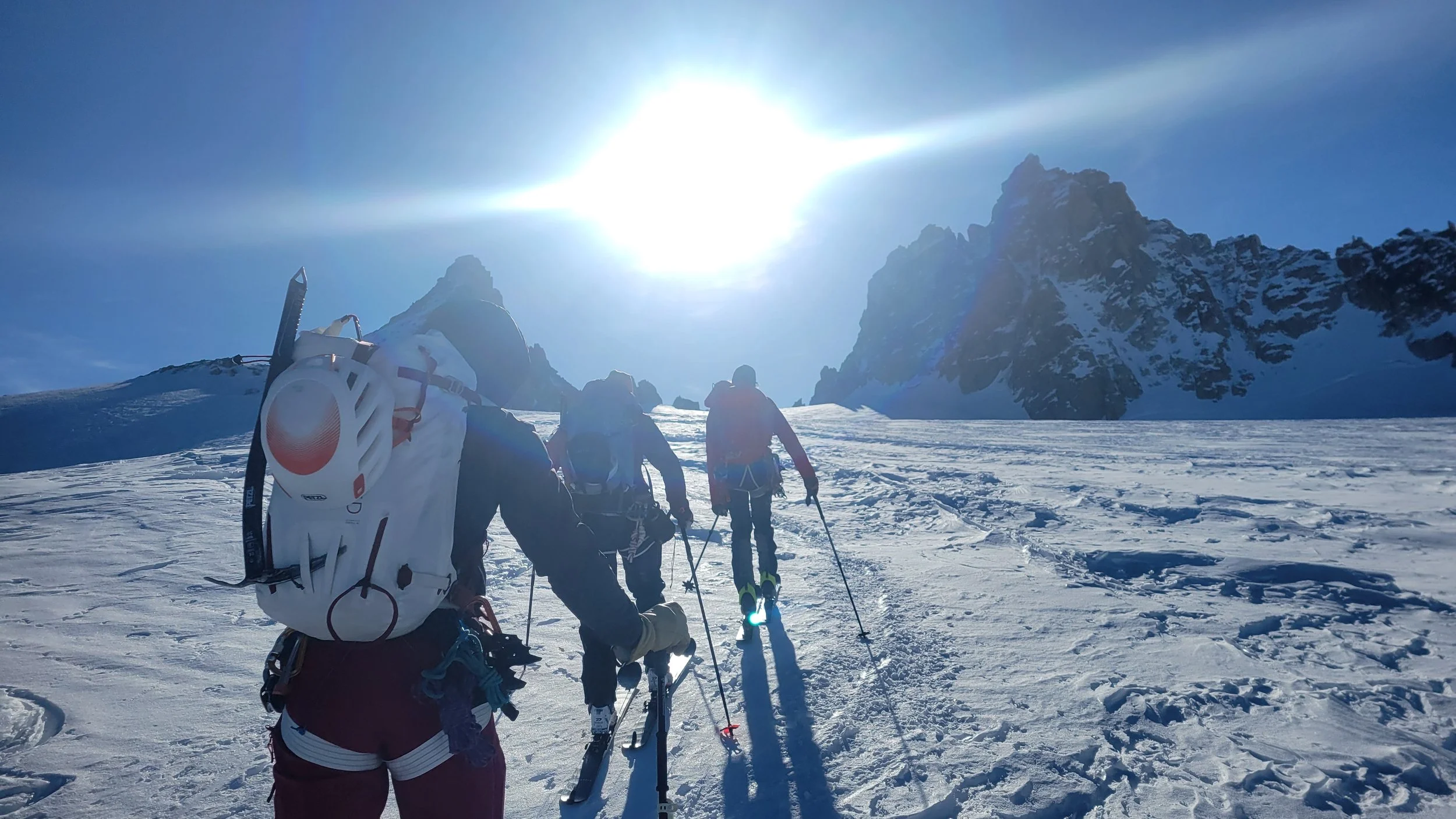 Vier Bergsteiger tragen Ausrüstung und gehen in einer verschneiten Berglandschaft bei Sonnenschein auf einer Gletscher. Schnee bedeckt den Boden und im Hintergrund sind steile, schneebedeckte Berge sichtbar.
