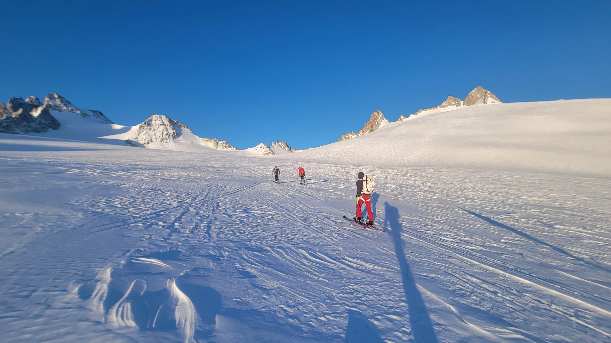 Drei Personen beim Skitouren in verschneiter Berglandschaft bei Sonnenuntergang, in den Bergen mit schneebedeckten Gipfeln unter blauem Himmel.
