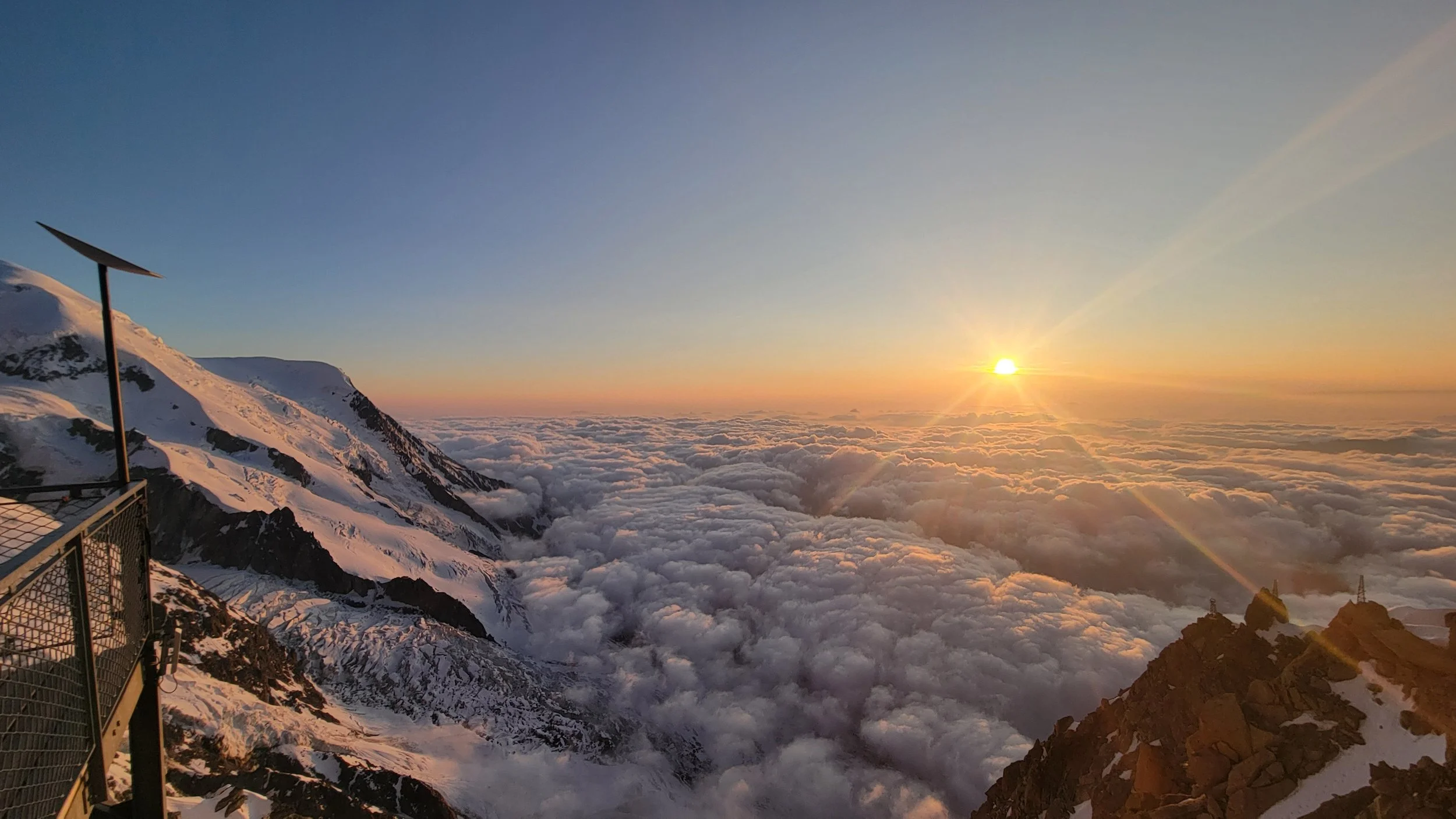 Blick von einem Berggipfel mit Schnee auf eine Wolkendecke bei Sonnenuntergang, mit einem Windrad im Vordergrund und einer bergigen Landschaft im Hintergrund.