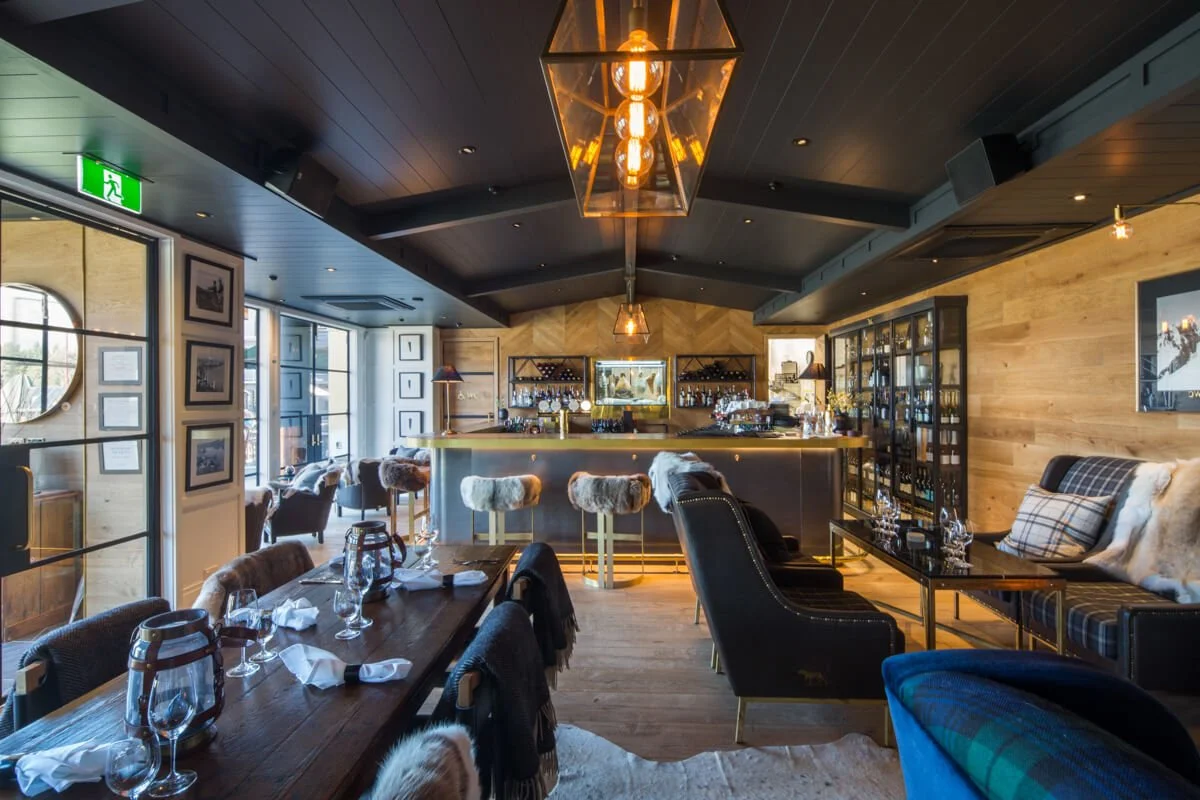Interior of a cozy, upscale restaurant or bar with a wood-paneled wall, a bar counter with bar stools, tables set with glassware and napkins, cushioned chairs, and decorative lighting.