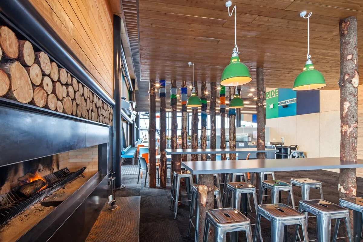 Interior of a modern restaurant or cafe with a wood-paneled ceiling, white tables, metal stools, and a fireplace with stacked logs. Wooden columns resembling tree trunks separate the seating area.