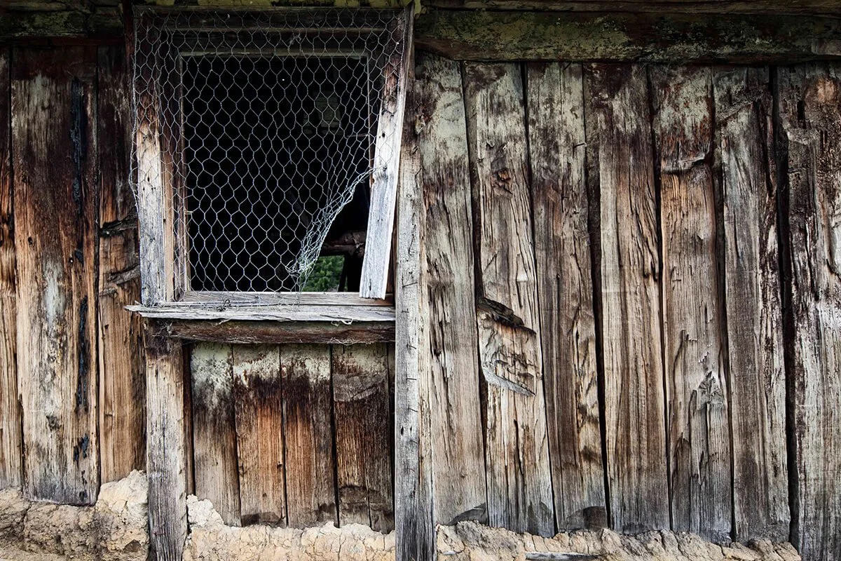An old, weathered wooden shed with a small window covered by a chain-link screen, set on a dirt ground.