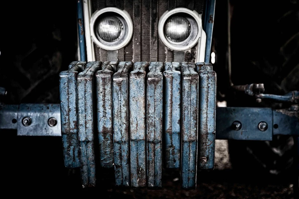 Close-up of a vintage car grille and front bumper with rusted and worn metal, featuring circular headlights in a dark setting.