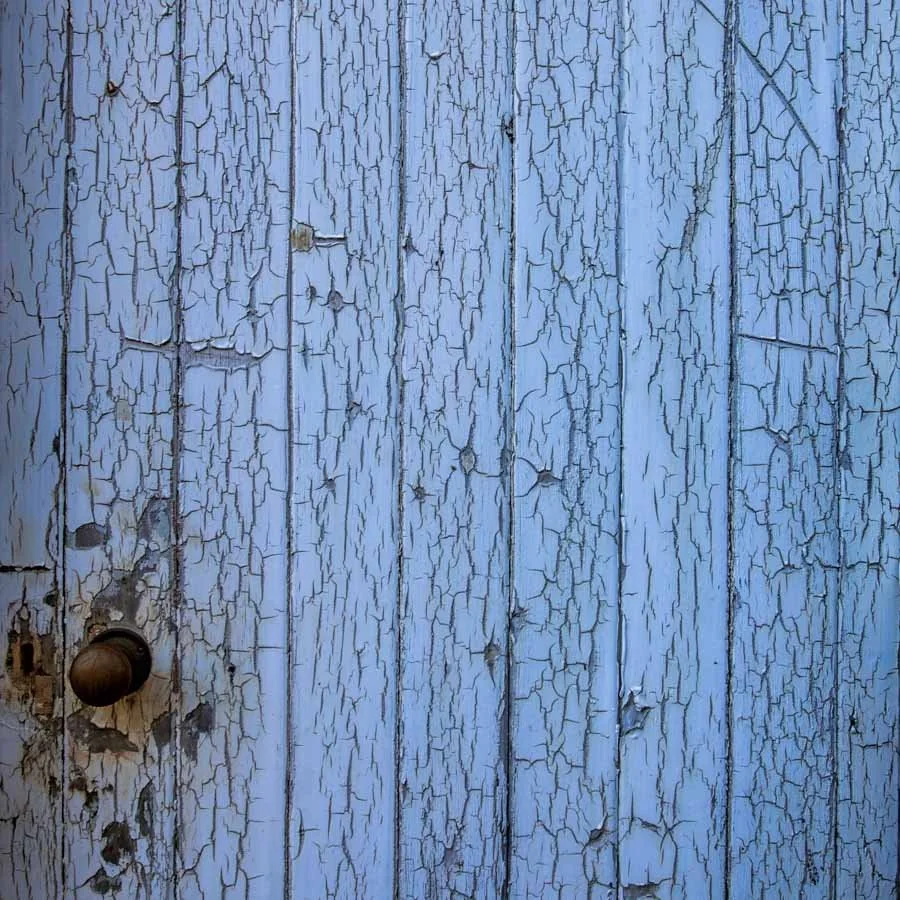Close-up of a weathered, light blue wooden door with peeling paint and a round doorknob.