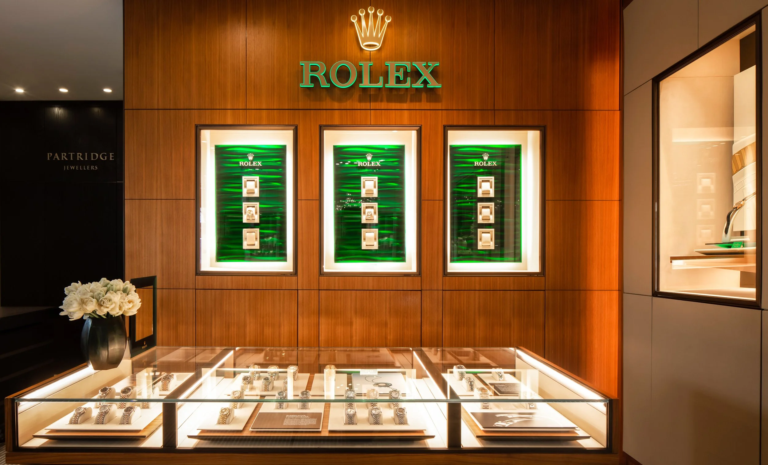 Display of luxury watches at a jewelry store with a prominent Rolex sign and logo on a wood-paneled wall, glass countertop with watches, and a vase with white flowers.