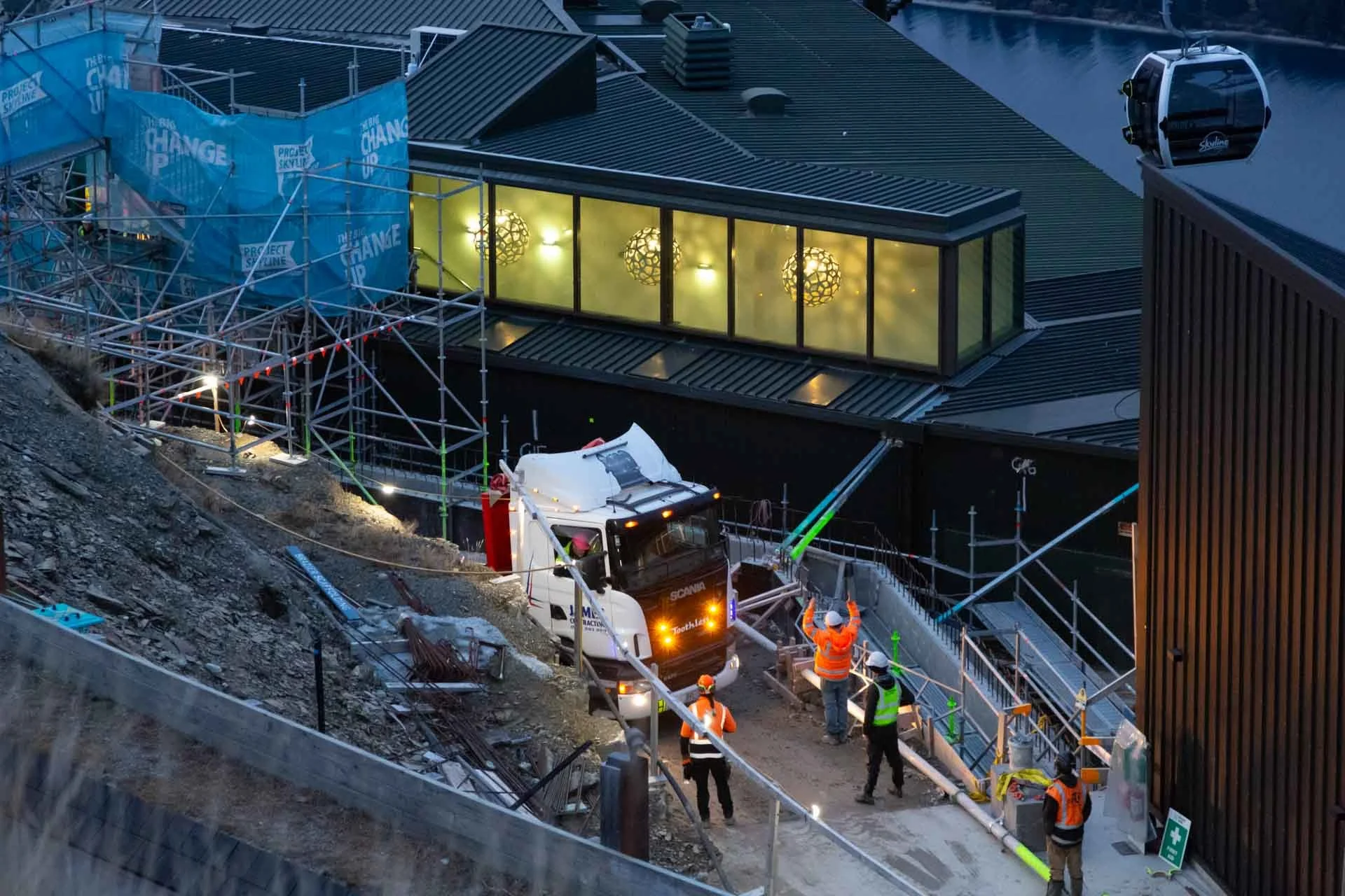 Construction site with workers in safety vests and helmets, a truck, scaffolding, and a building with large yellow windows at dusk.