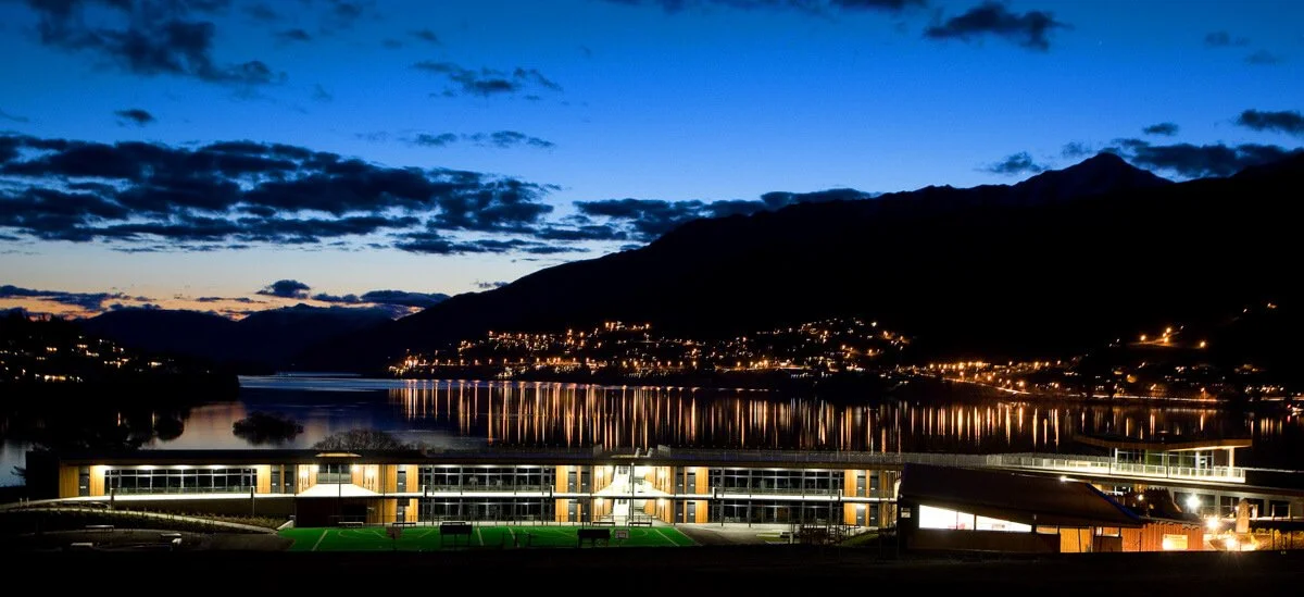 Nighttime view of a modern school building in front of a body of water with mountains and city lights in the background.