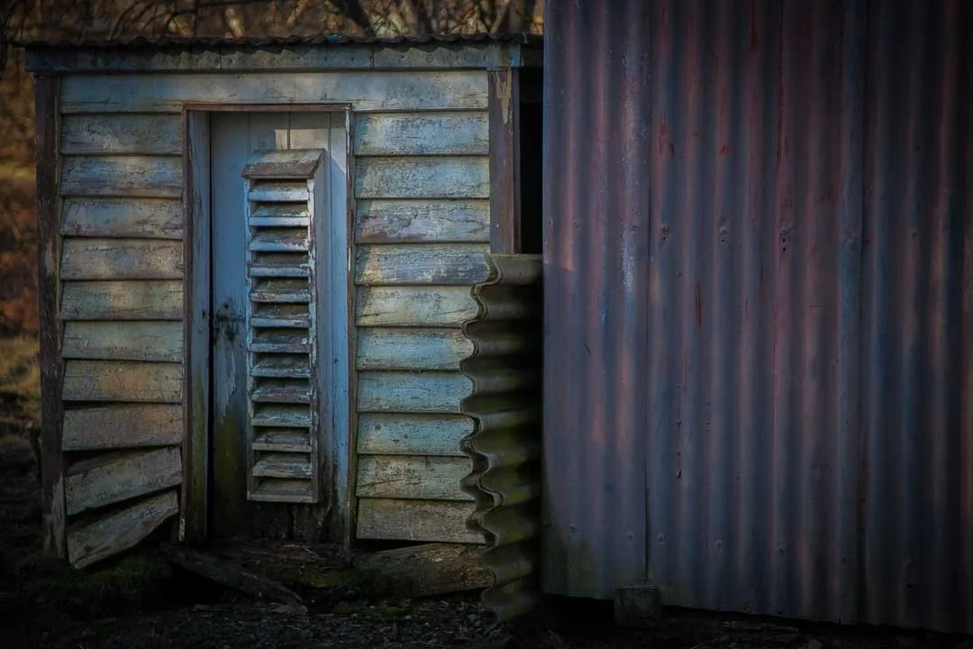 Close-up of an old weathered wooden and metal shed with a blue vented door and rusted corrugated metal siding.