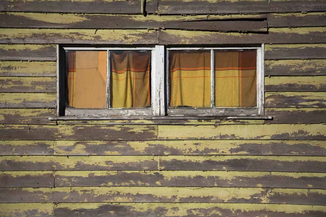 A weathered, yellow wooden house with peeling paint, featuring a small double window with white trim and curtains inside.
