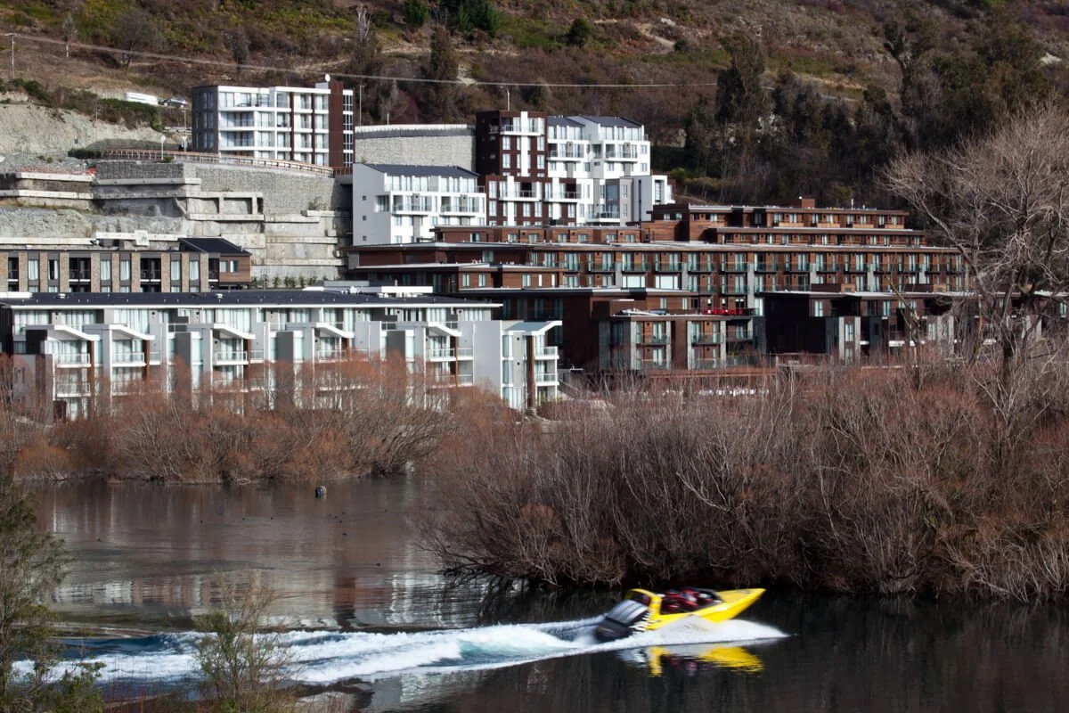 Modern apartment buildings on a hillside behind a river, with a yellow speedboat speeding across the water in the foreground.