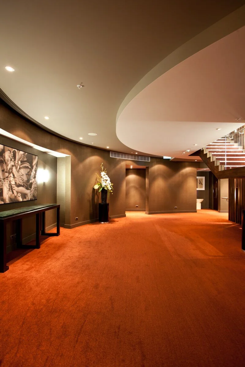 Interior of a modern, stylish building with a curved ceiling, reddish carpet, black tables, and a flower vase on a pedestal.