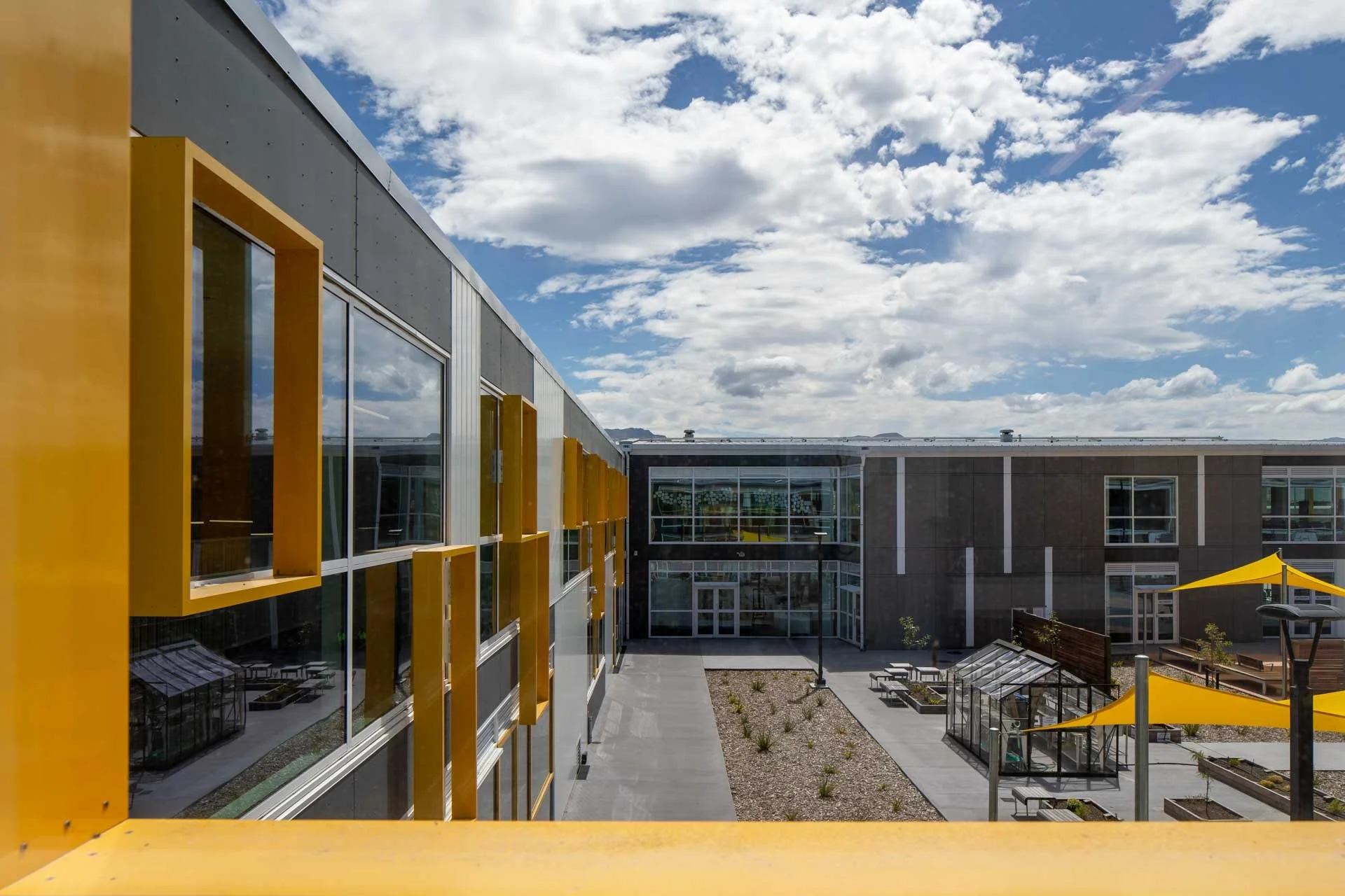 Modern building with large windows and yellow window frames, outdoor courtyard with gravel and small plants, glass greenhouses, and yellow shade sails under a partly cloudy sky.