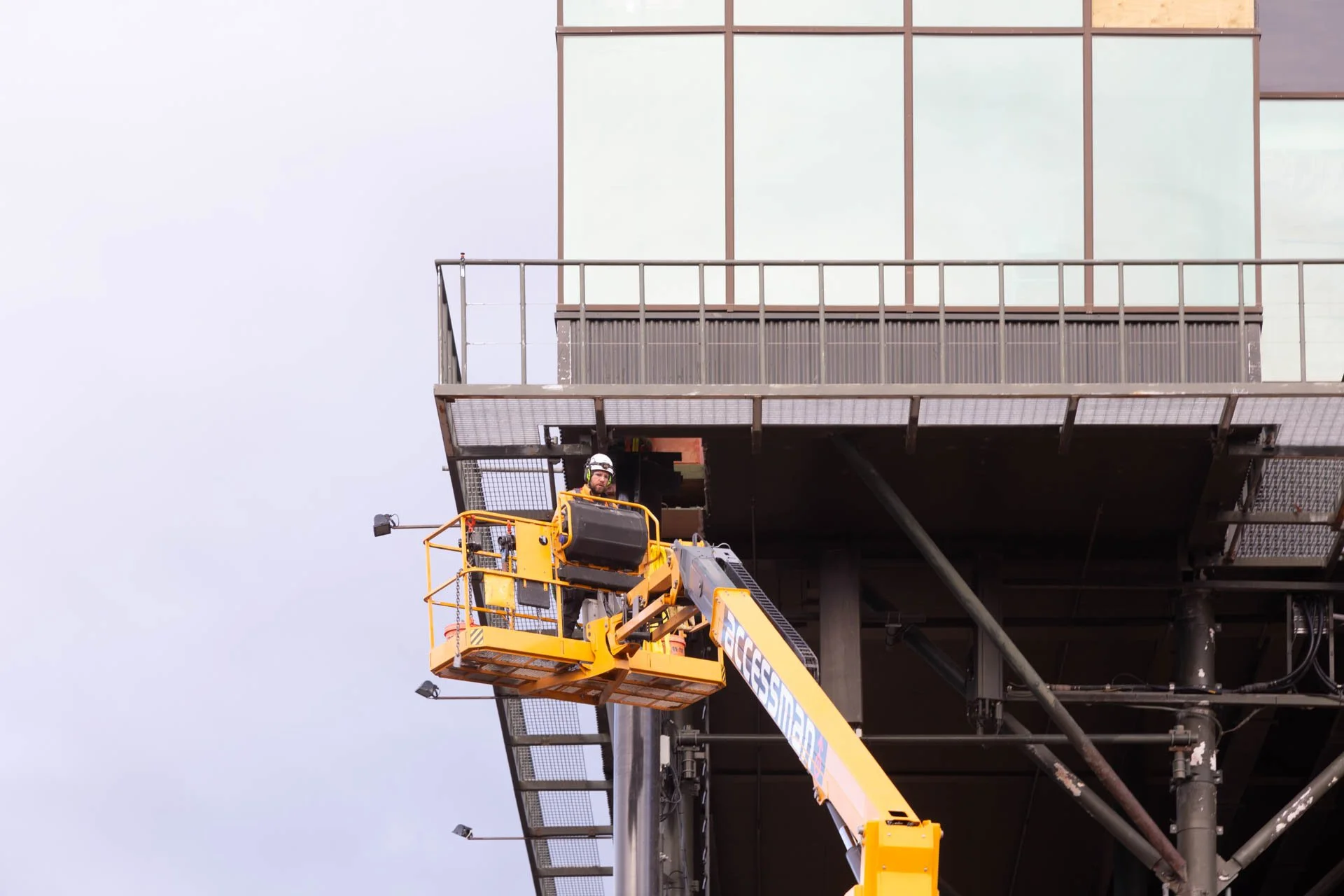 A construction worker wearing a helmet stands in a yellow lift basket on a tall scaffold, working on the exterior of a building with glass windows.