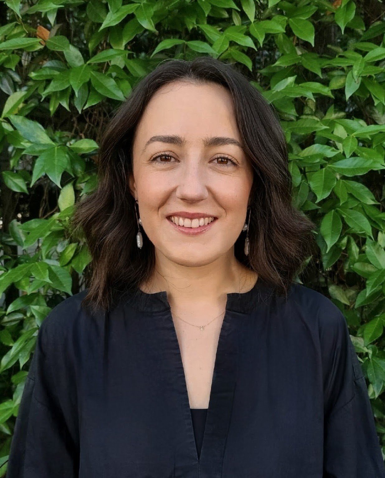 A woman with shoulder-length dark brown hair, smiling, wearing dangle earrings and a black blouse, standing in front of green leafy bushes.