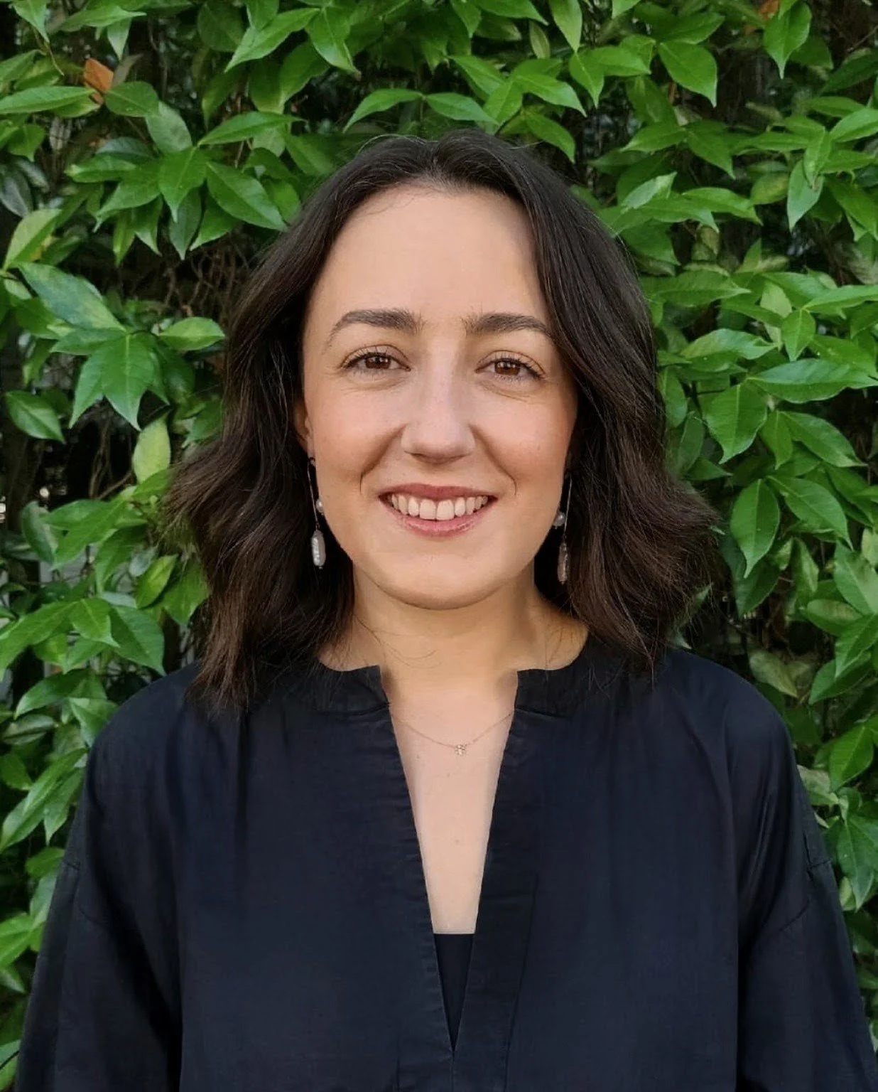 A woman with shoulder-length dark brown curly hair smiling in front of green leafy background, wearing a black top and pearl earrings.