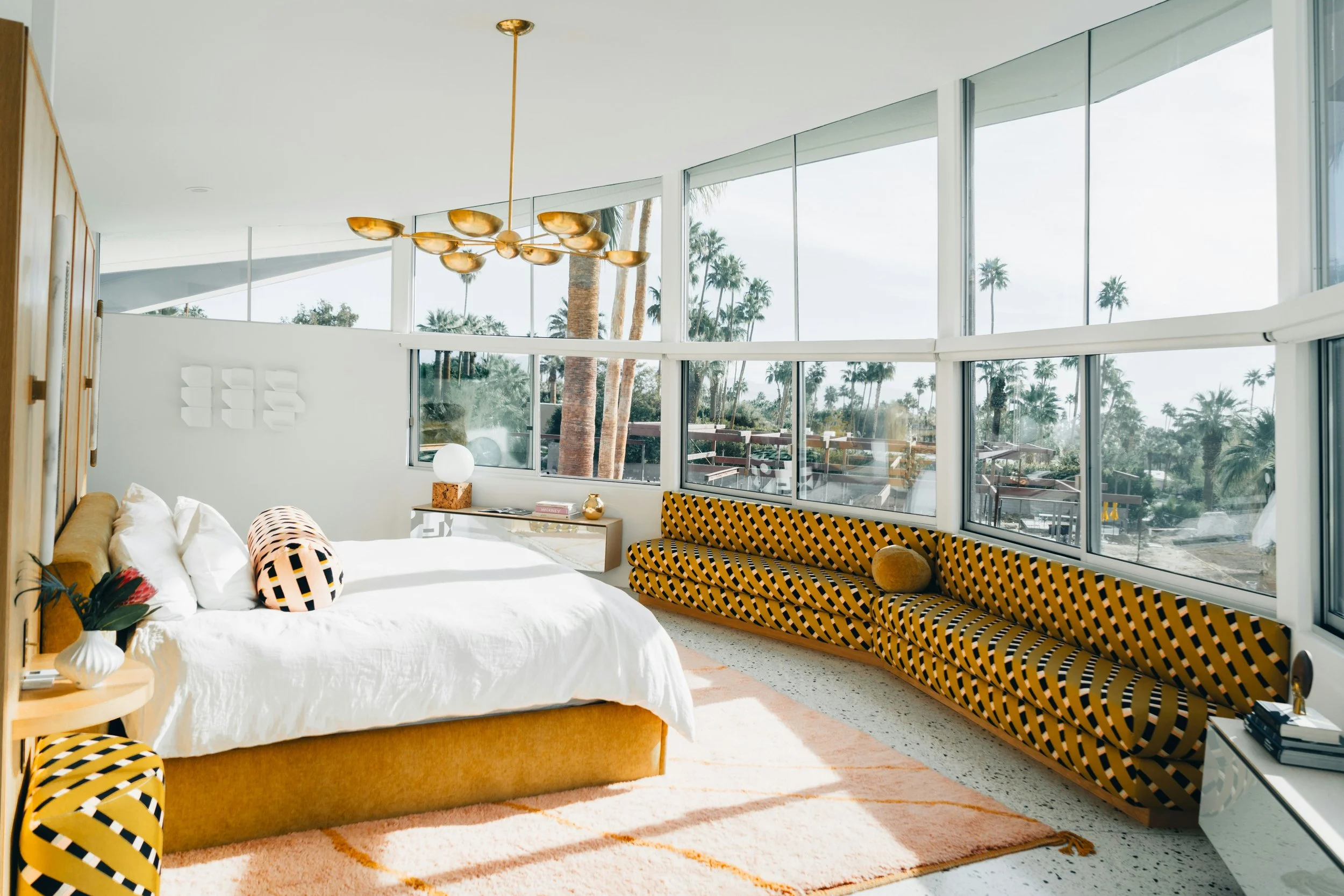 Modern bedroom with large windows, white bed with yellow upholstered base, geometric-patterned yellow and black sofa, side tables with decorative items, pendant lighting, and tropical outdoor scenery with palm trees