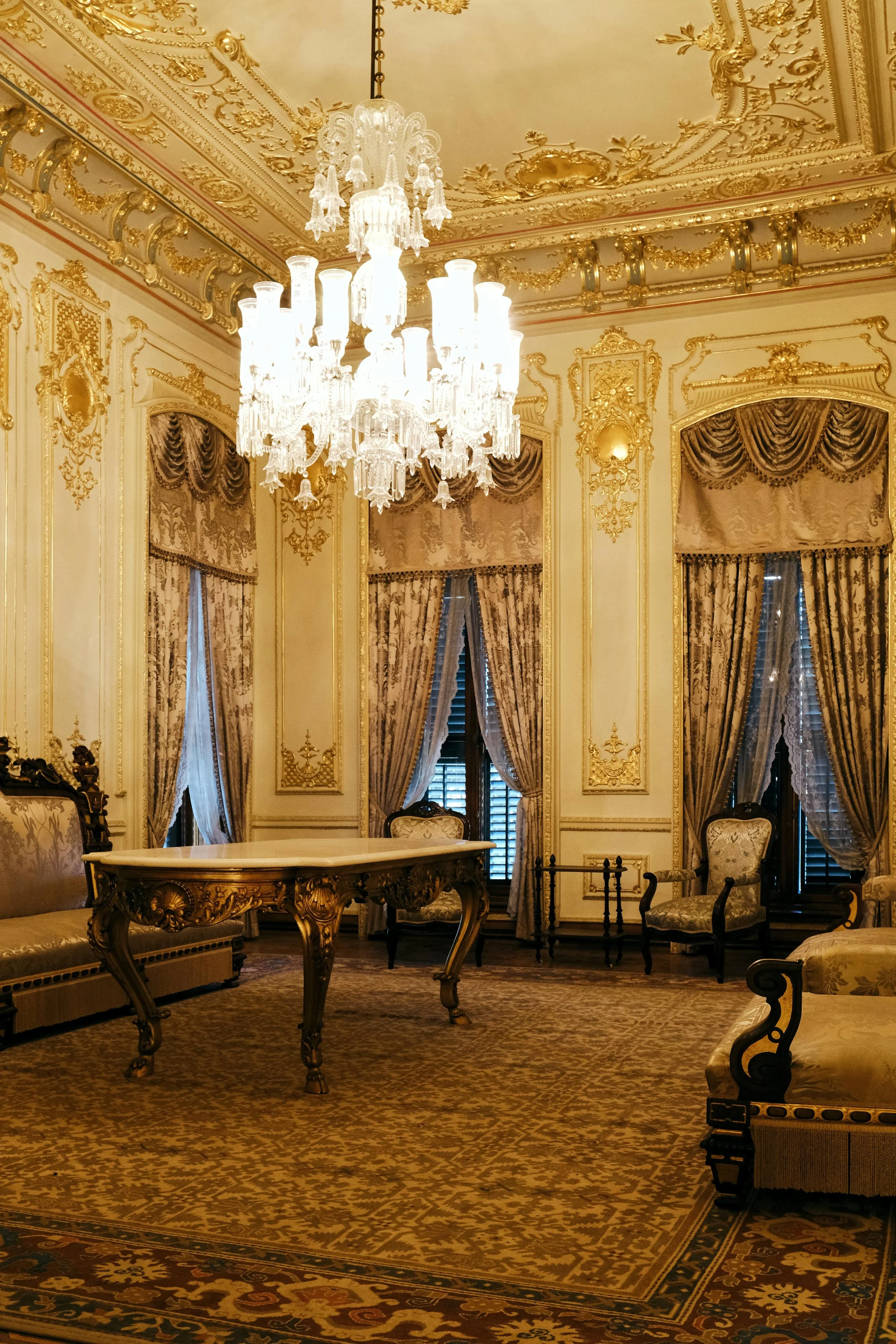 Luxurious, ornate room with gold detailing, a large chandelier, heavy drapes, vintage furniture, and patterned carpet.