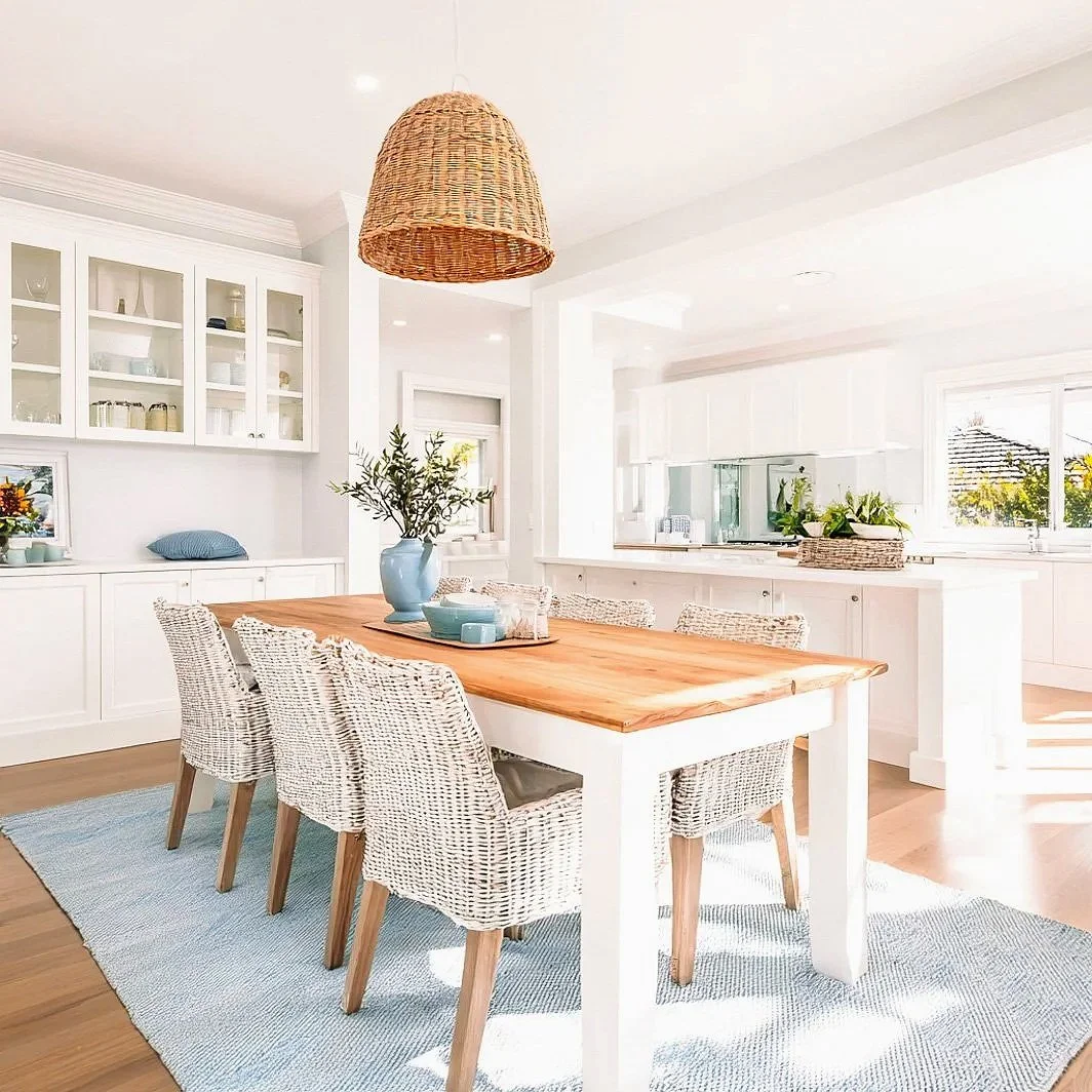 Bright, airy dining room with a wooden table, six woven chairs, a blue vase with greenery, white cabinets, and large windows