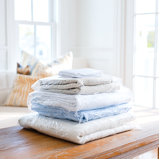 Stack of folded bedding and linens on a wooden table in a bright room with large windows.