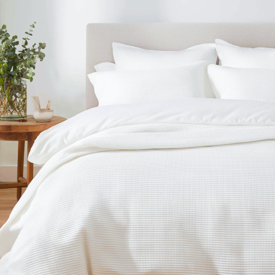 A neatly made bed with white bedding and four white pillows, next to a wooden nightstand with a plant and a small cup or container.