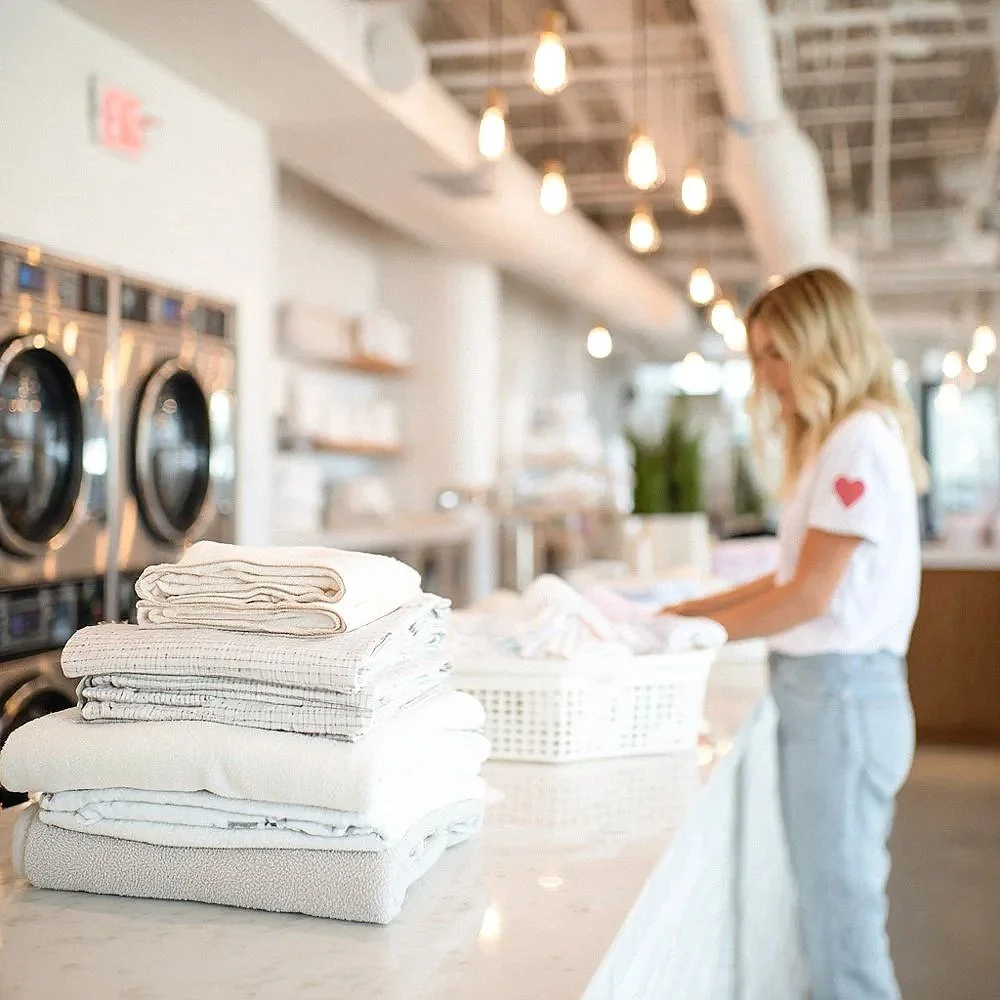 A woman folding laundry at a counter with stacks of white linens in a modern laundromat with hanging lightbulbs.