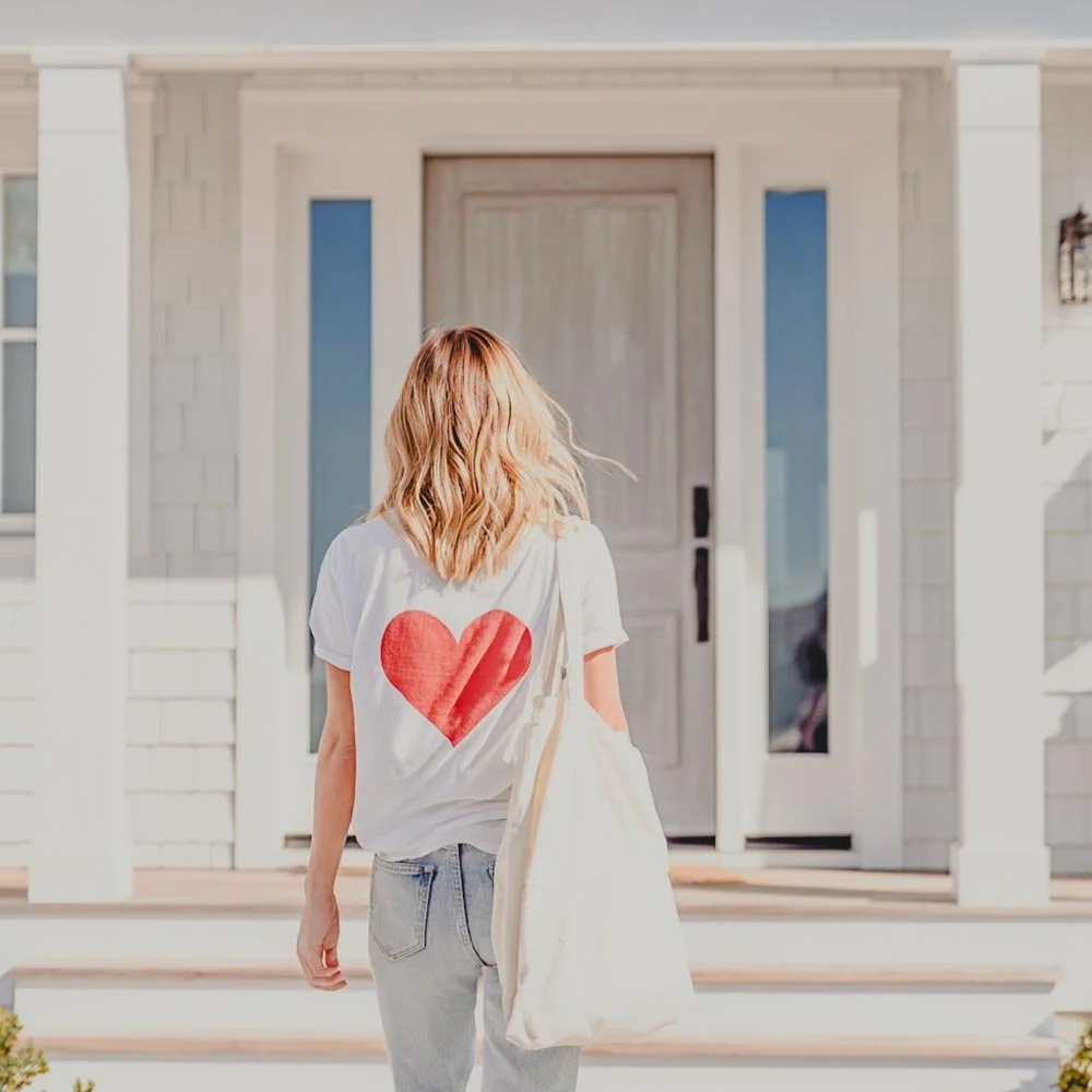 A woman with shoulder-length blonde hair walking towards a white house. She is wearing a white T-shirt with a red heart on the back and carrying a beige bag.
