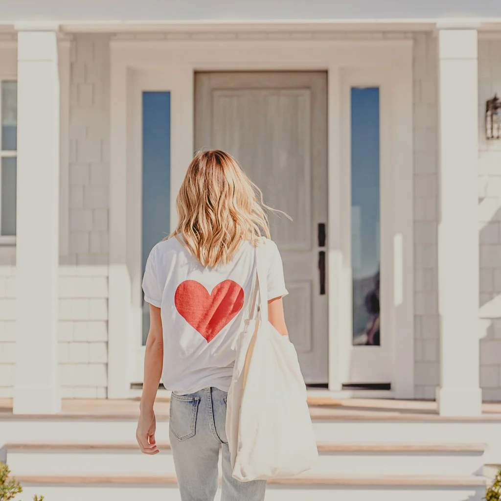 A woman with blonde hair wearing a white T-shirt with a red heart on the back, jeans, and carrying a large tote bag, walking towards the front door of a house.