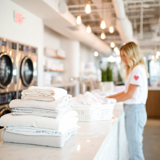 A woman folding laundry in a modern laundromat with laundry stacks on a marble counter in the foreground.