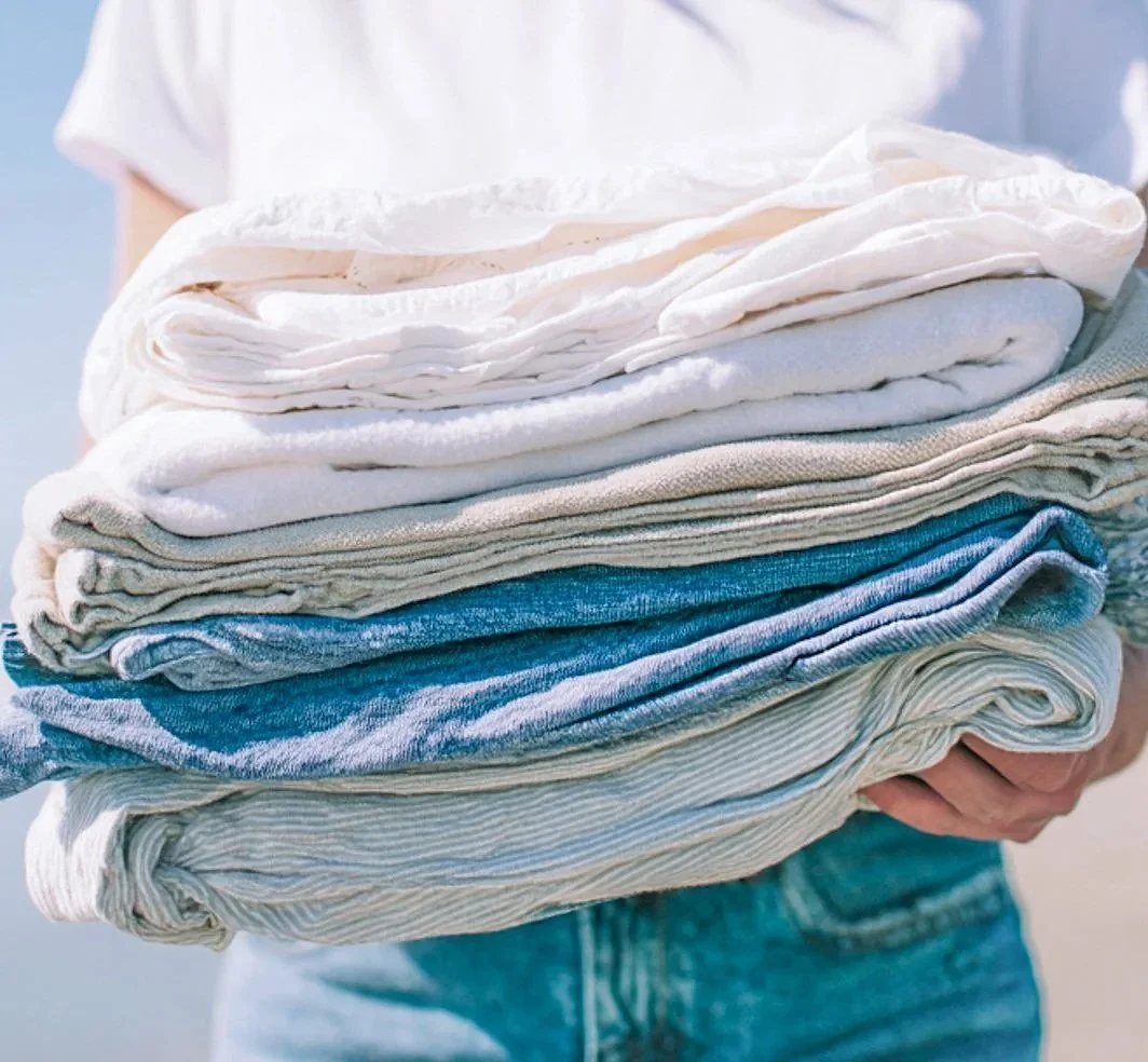 A person holding a large stack of folded clothes, including white, beige, and blue garments.