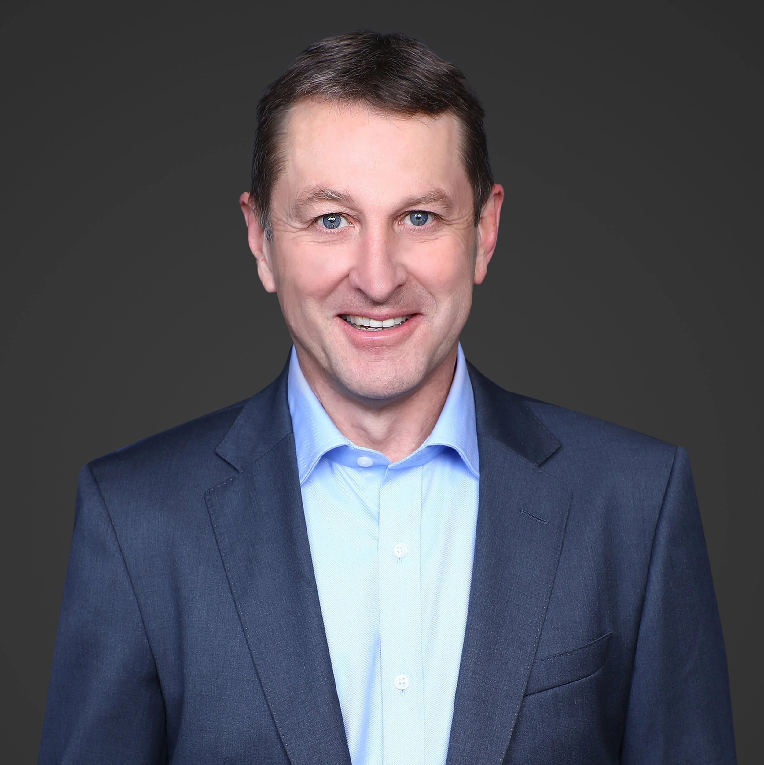 Close-up of a middle-aged man with short, dark brown hair and blue eyes, wearing a navy blue suit and a light blue shirt, smiling against a plain dark gray background.