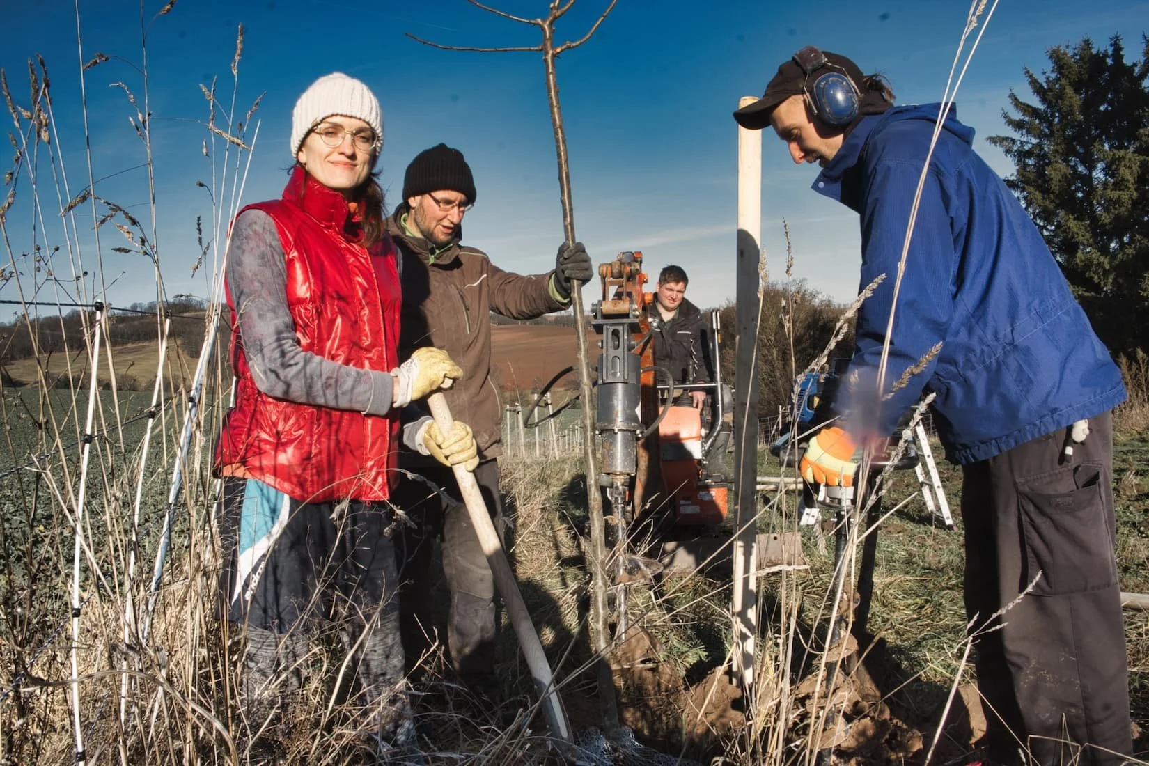Vier Personen beim Baum-Pflanzprojekt im Freien, eines mit roten Jacke, einer mit brauner Jacke, einer mit blauer Jacke mit Kopfhörer, alle arbeiten mit Werkzeugen und Maschinen in einem landwirtschaftlichen Feld, sonniger Tag.