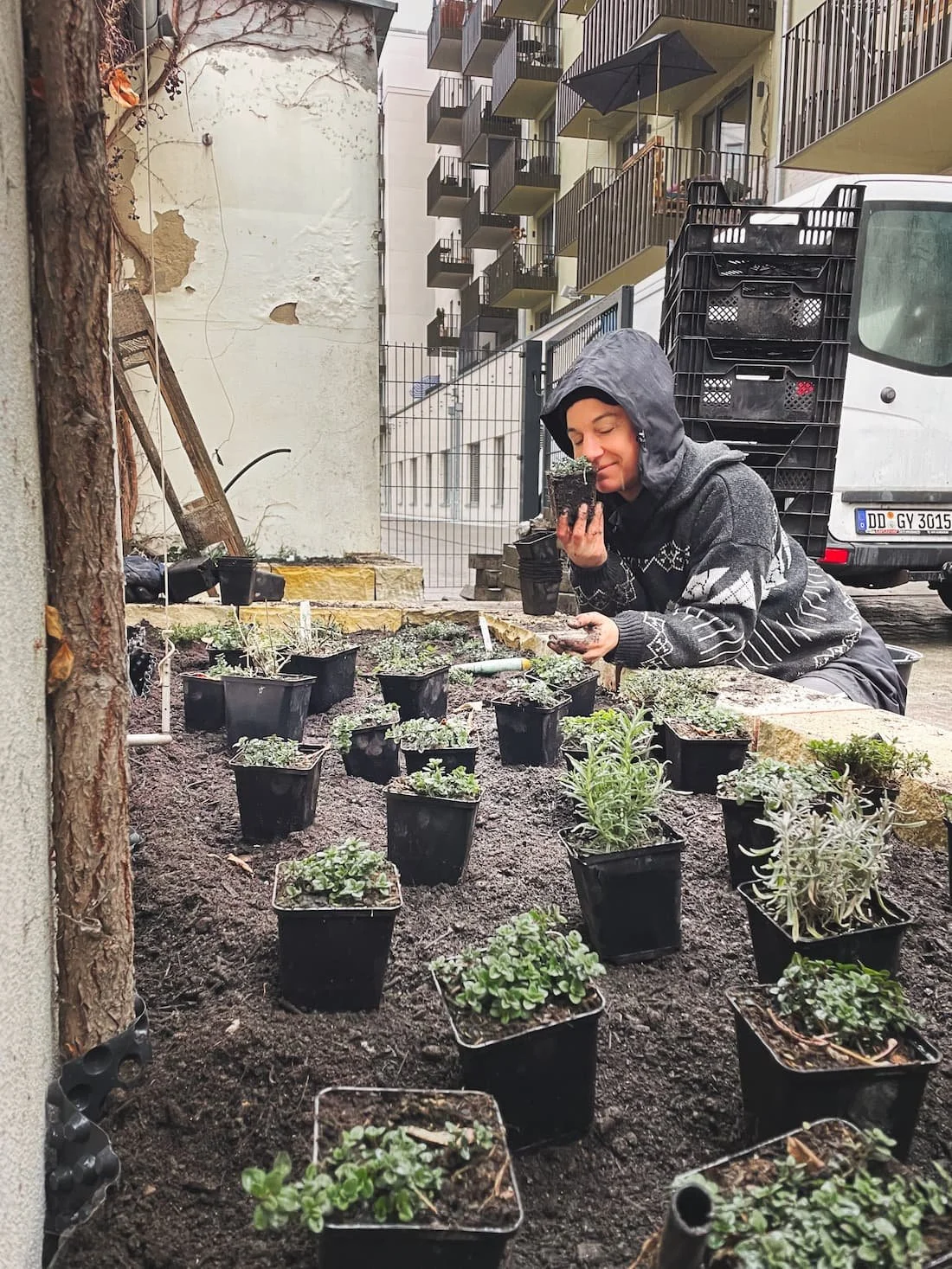 Eine Person in einem grauen Hoodie, die auf einem Baugrundstück auf dem Boden kniet, während sie Pflanzen in kleine schwarze Töpfe setzt. Im Hintergrund ist ein weißer LKW und ein mehrstöckiges Wohngebäude mit Balkonen zu sehen.