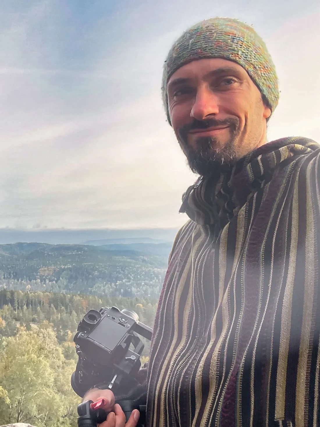 Mann trägt bunte Mütze, hält Kamera, mit Blick auf Landschaft aus Wäldern und Hügeln bei bewölktem Himmel