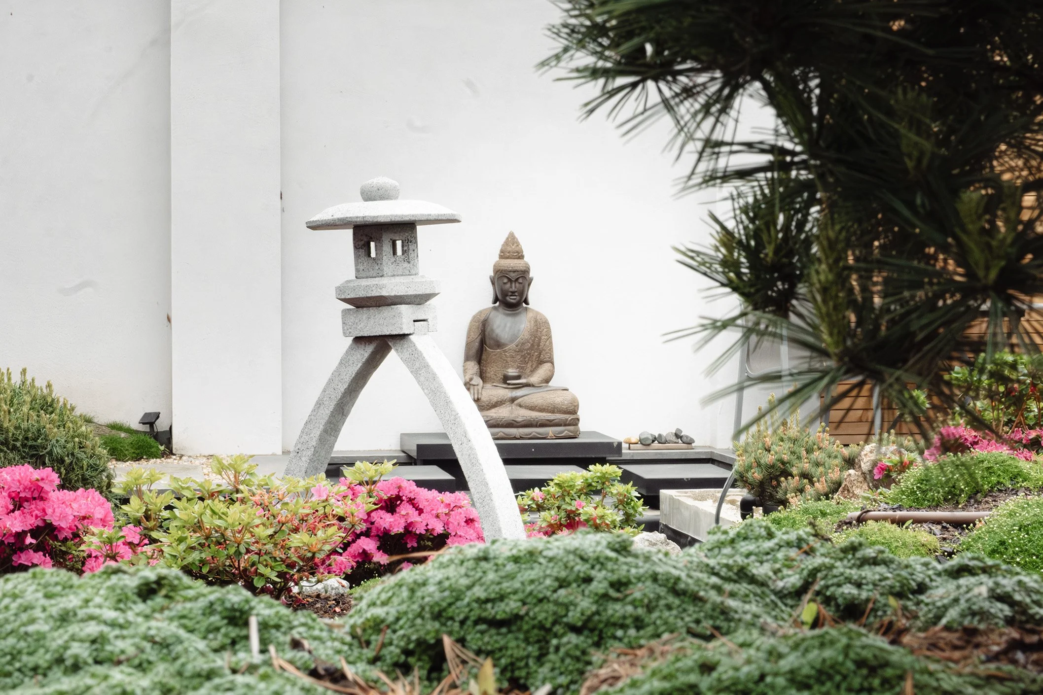 Garten mit Skulpturen eines Buddha und einer japanischen Laterne, umgeben von blühenden Pflanzen und grünem Moos.