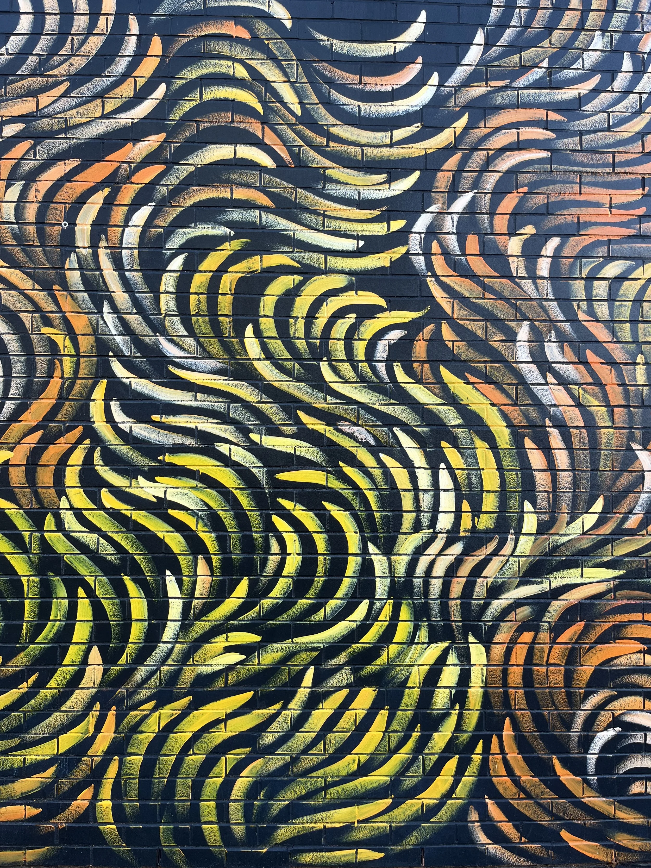 Colorful abstract mural with swirling curved lines in shades of yellow, orange, white, and green on a black brick wall.