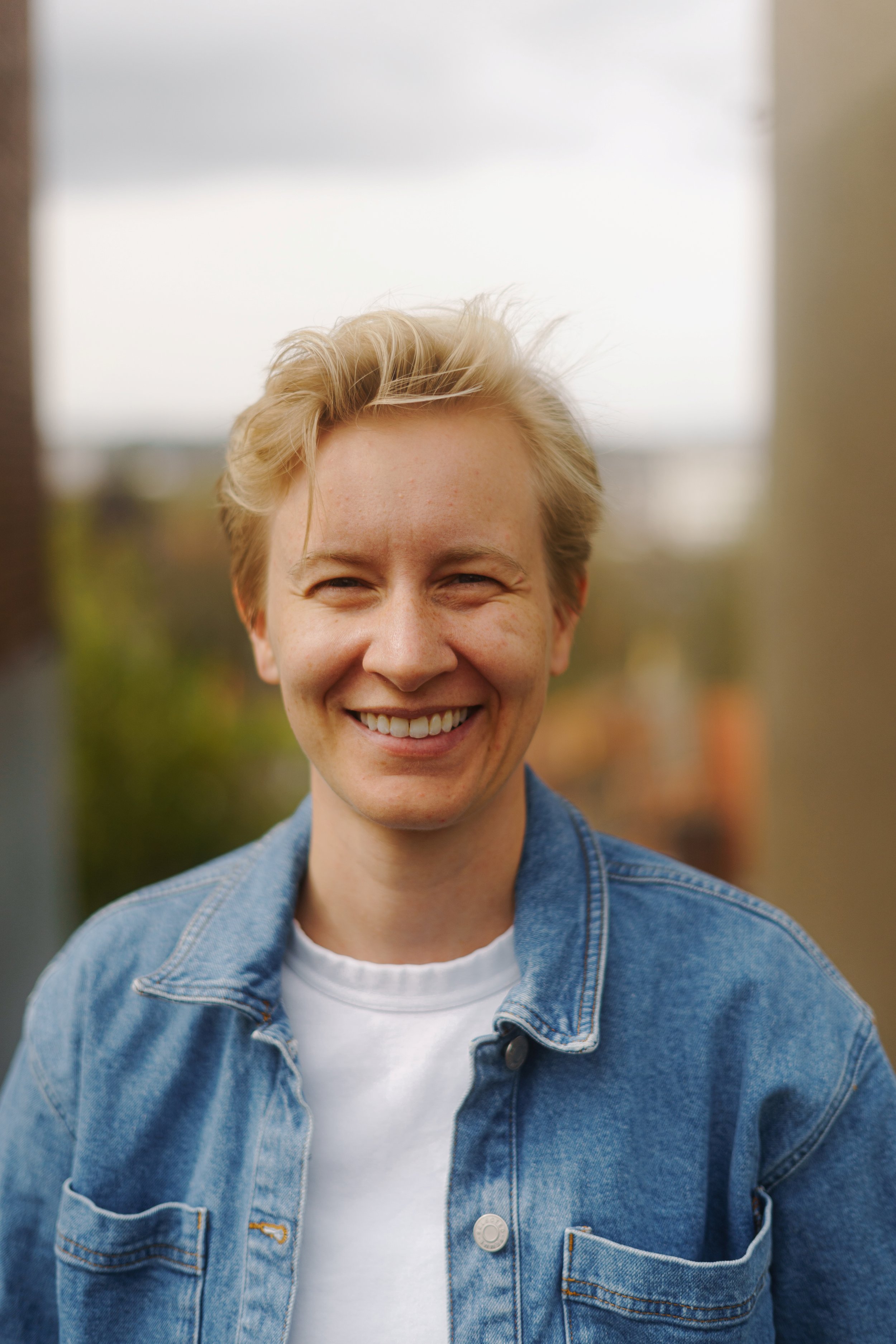 A smiling person with short, blonde hair wearing a white t-shirt and a denim jacket, standing outdoors with blurred background.